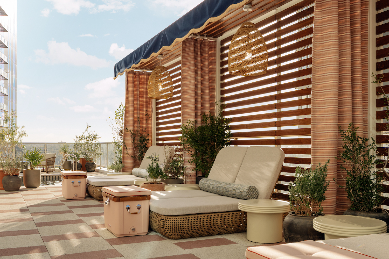 A modern outdoor lounge on the Paseo features cushioned wicker chairs, small tables, potted plants, and hanging woven lamps under a blue-striped awning. Slatted wood panels offer partial shade beneath a bright, clear sky.