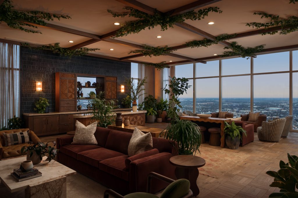 Elegant living room with large windows showcasing a city view along the Paseo, filled with green plants, brown and rust-colored sofas, wooden furniture, and ceiling beams adorned with greenery. Warm lighting creates a cozy, inviting atmosphere.