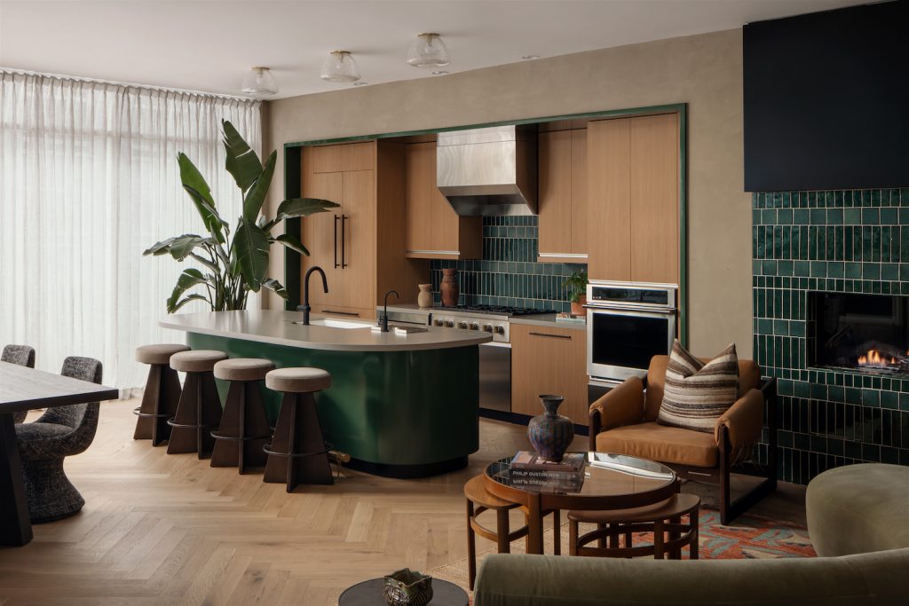 Modern kitchen with a curved island, green base, and four bar stools. Wood cabinets, green tile backsplash, built-in oven, and fireplace with matching Paseo green tile. Large plant and cozy seating add warmth to the space.