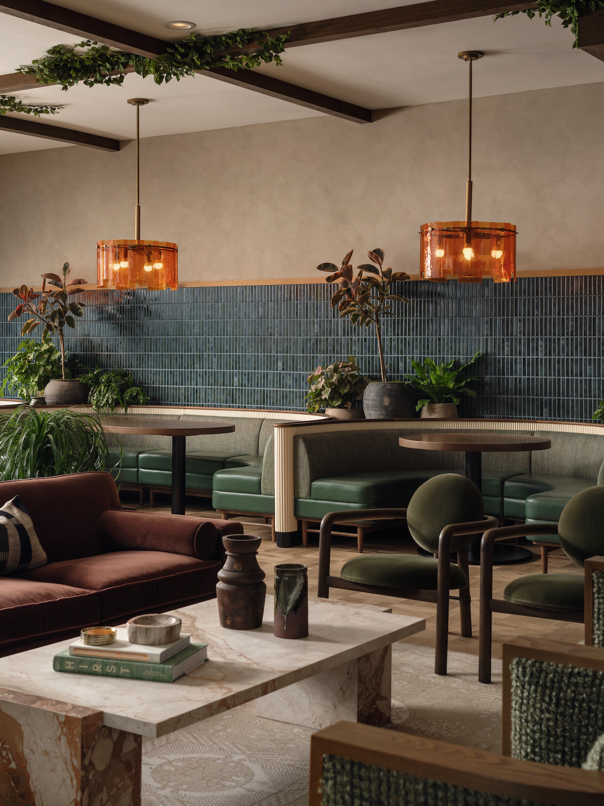 A stylish lounge at Paseo with green booths, brown armchairs, a marble coffee table, and indoor plants. Amber pendant lights hang from the ceiling above seating, and a blue tiled wall lines the back.