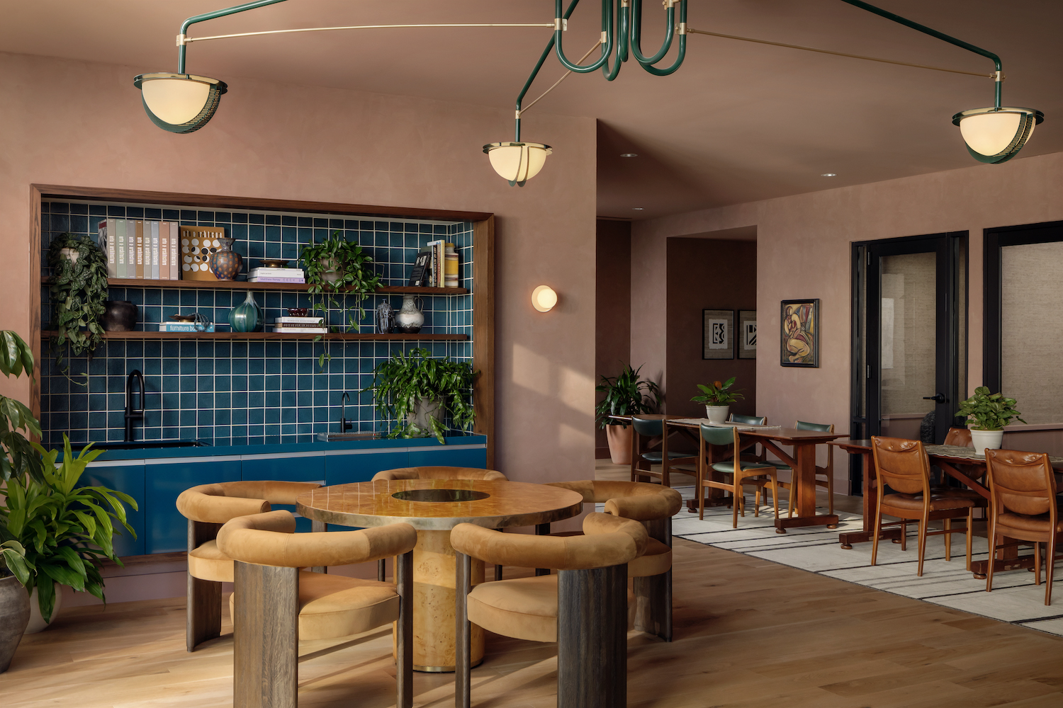 A modern Paseo-inspired room with tan walls, round wooden tables with cushioned chairs, a blue tiled kitchenette with shelves and plants, and a seating area with brown chairs and framed art on the wall. Contemporary lighting hangs above.