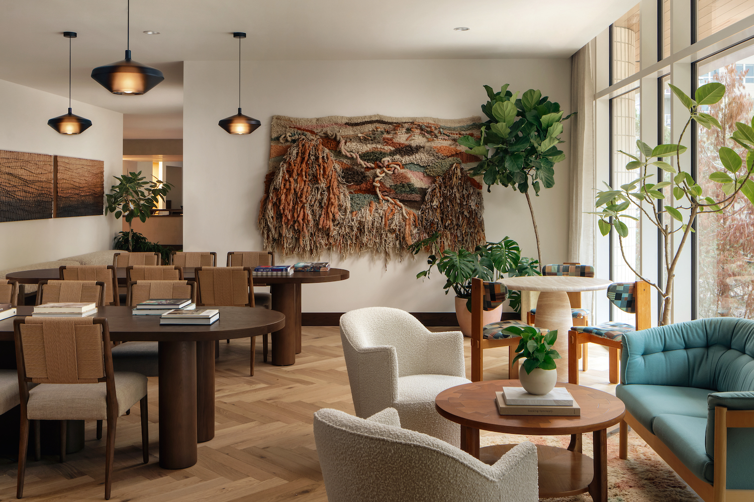 A cozy, modern lounge with round wooden tables, cushioned chairs, and green plants awaits at Paseo. Large windows, textured wall art, and pendant lights add warmth to the neutral-toned, inviting space.