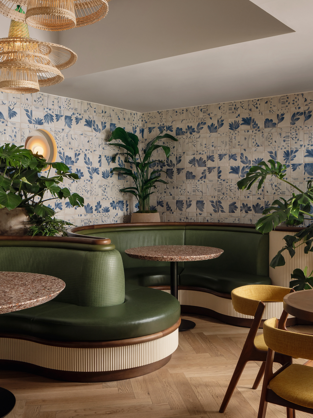 A cozy Paseo restaurant corner with green curved banquette seating, round marble tables, yellow chairs, leafy potted plants, and blue floral-patterned wallpaper, all softly illuminated by woven pendant lights.