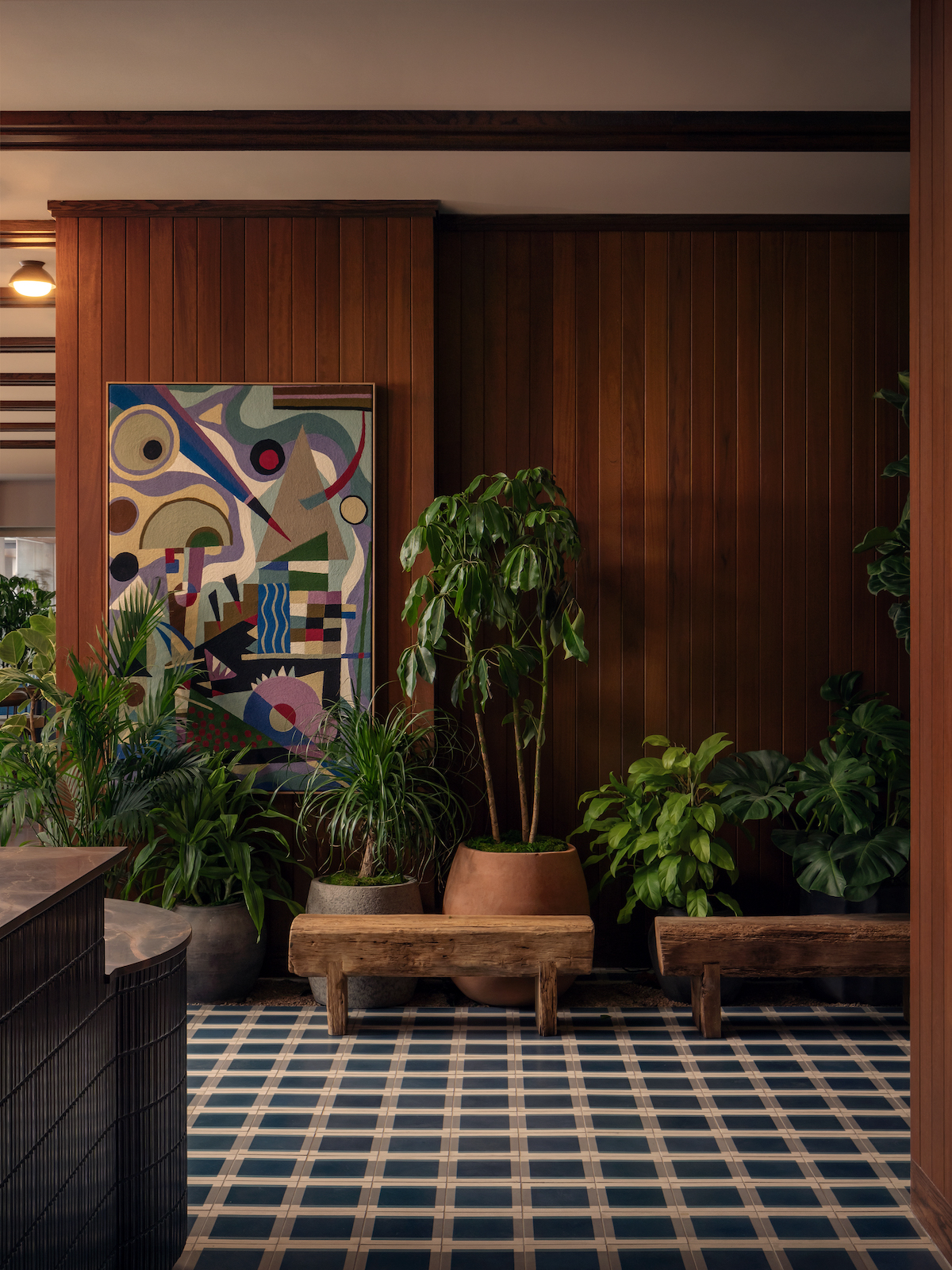 A cozy room with wood-paneled walls, potted green plants on wooden benches, a geometric patterned tile floor reminiscent of a Paseo, and a colorful abstract painting hanging on the wall.