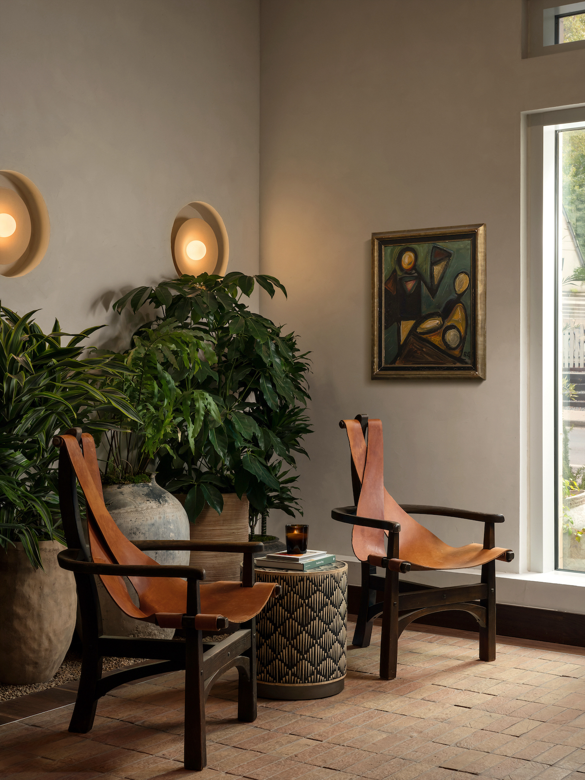 Two leather chairs with wooden frames face a small round table holding a drink, set on a brick floor near large potted plants. A modern painting, wall sconces, and a tall window create a cozy, stylish Paseo-inspired room.