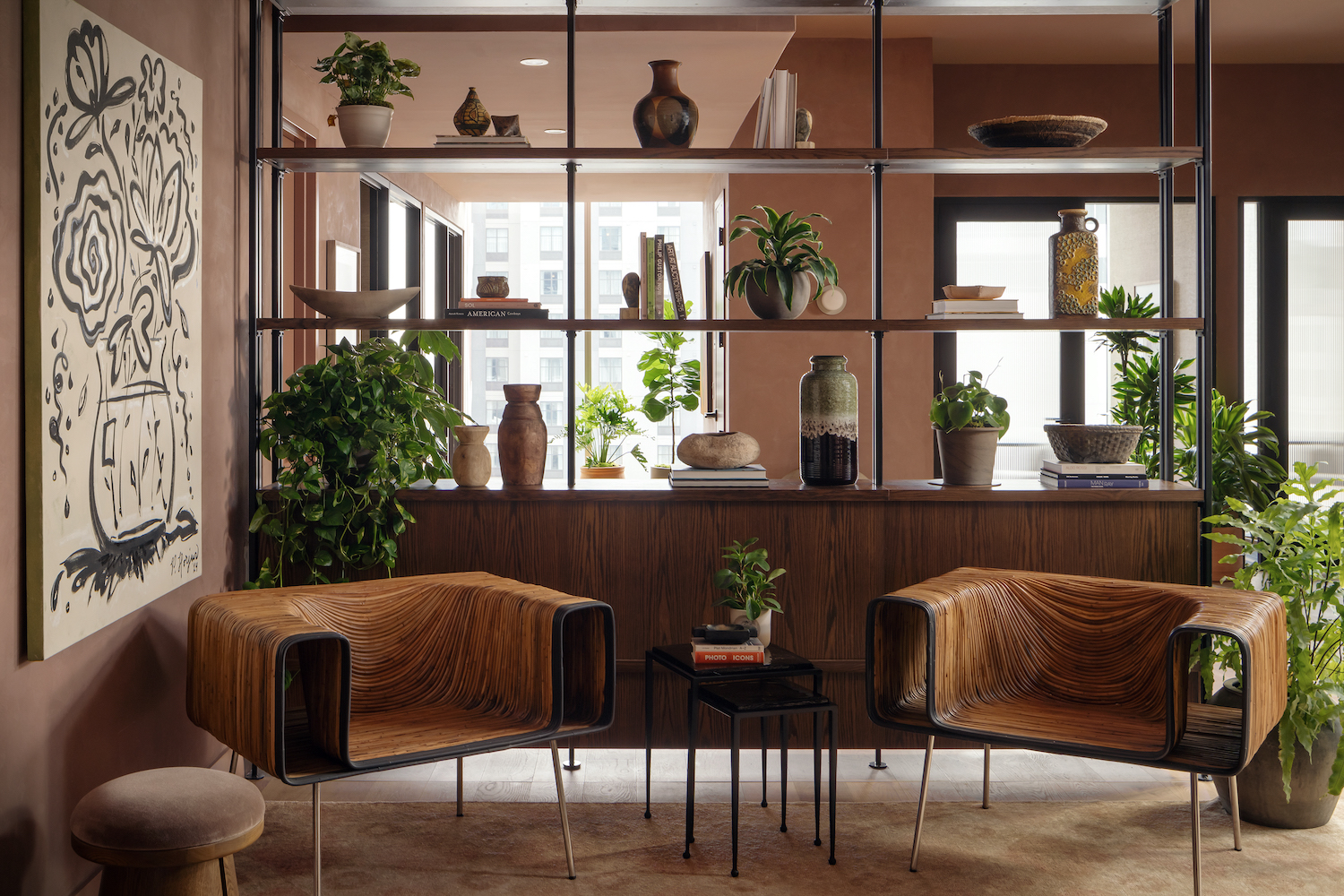 A cozy modern lounge with two wooden armchairs, small tables, potted plants, and open shelves holding pottery and decor creates a relaxing Paseo vibe. Sunlight streams in behind the shelves, highlighting a large abstract floral painting on a brown wall.