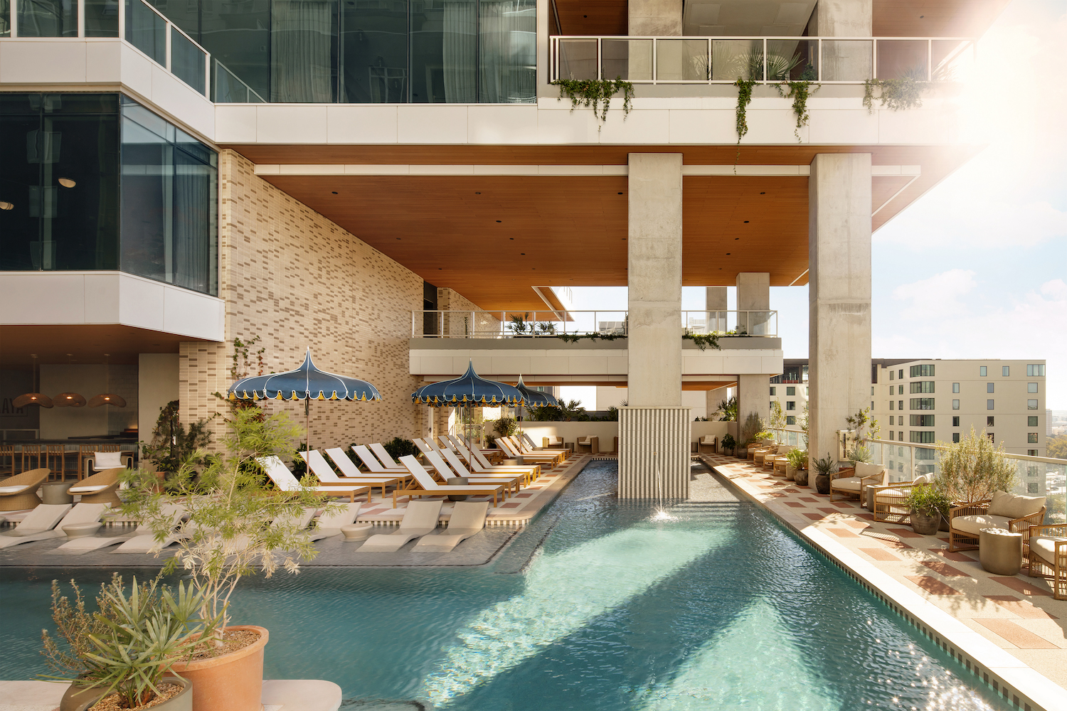 A modern rooftop pool with lounge chairs, blue-striped umbrellas, and lush plants creates a luxurious paseo retreat against a high-rise building. Sunlight streams in, enhancing the relaxing atmosphere.