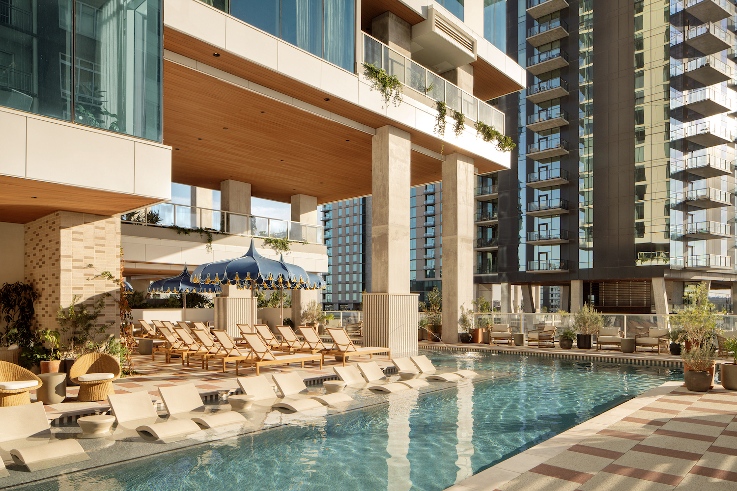 A modern outdoor pool along the Paseo features lounge chairs, built-in loungers in the water, and is surrounded by tall glass buildings. A canopy shades a seating area, while potted plants add greenery to this bright, inviting scene.