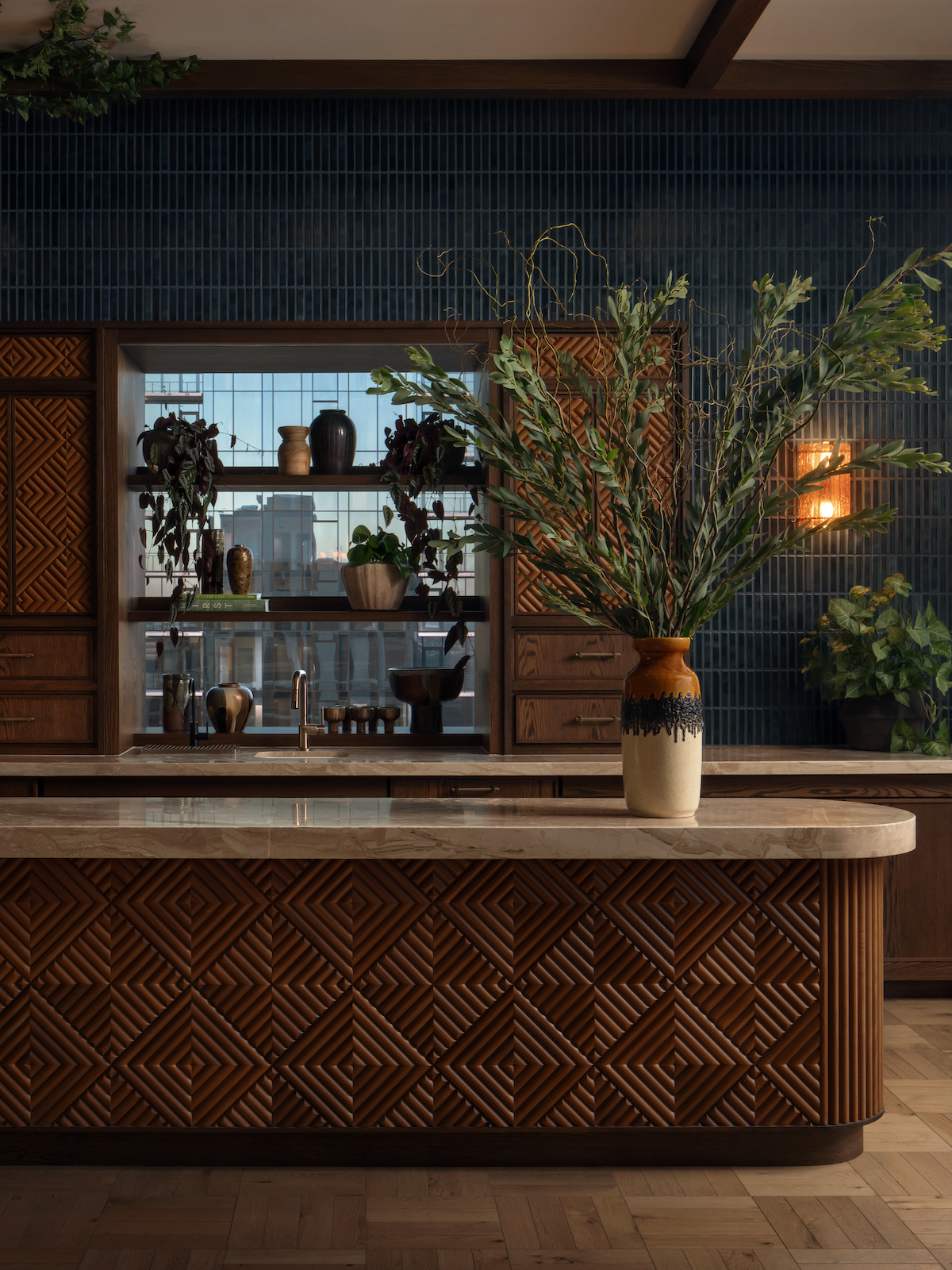 A stylish kitchen by Paseo features a marble island with geometric wood patterns, a tall vase with leafy branches, dark blue tiled backsplash, wooden cabinets, and potted plants near a window. Warm lighting creates a cozy atmosphere.