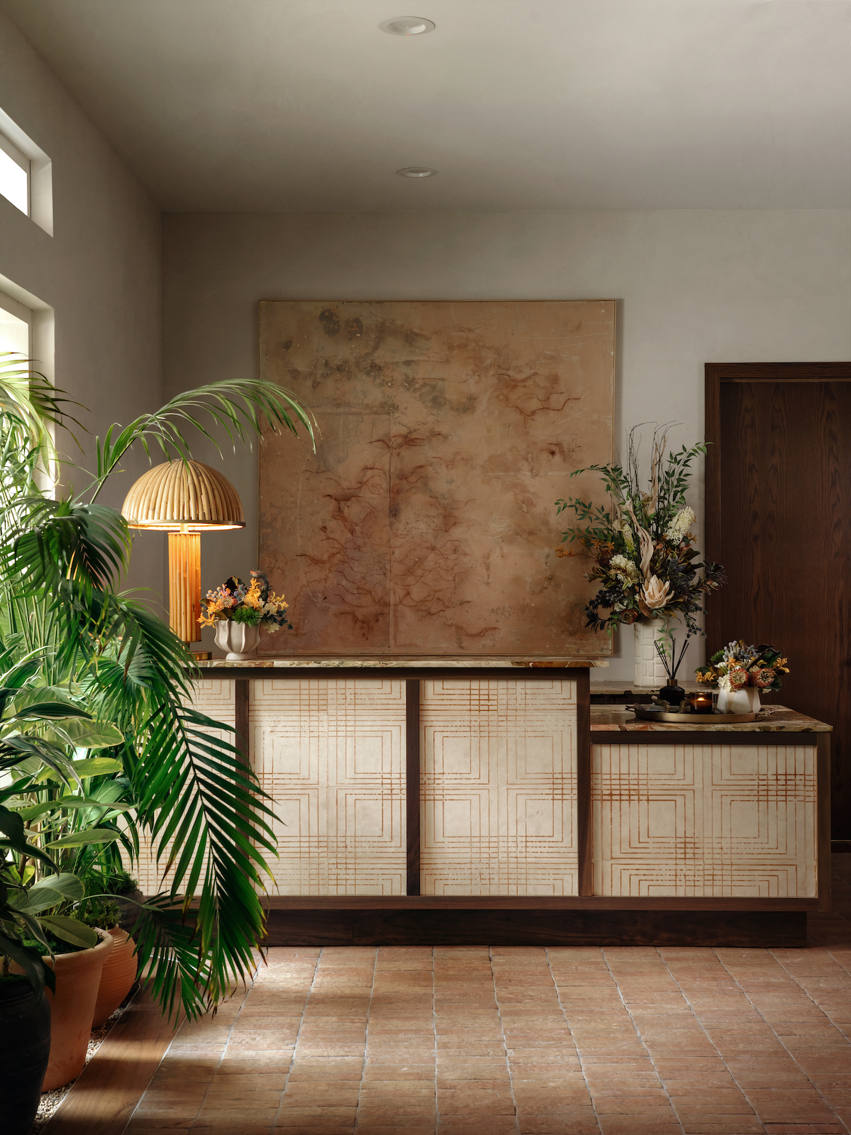 A stylish Paseo room features a patterned reception desk, large leafy plant, marble lamp, floral arrangements, and a large abstract artwork on the wall, with warm lighting and earthy tones throughout.
