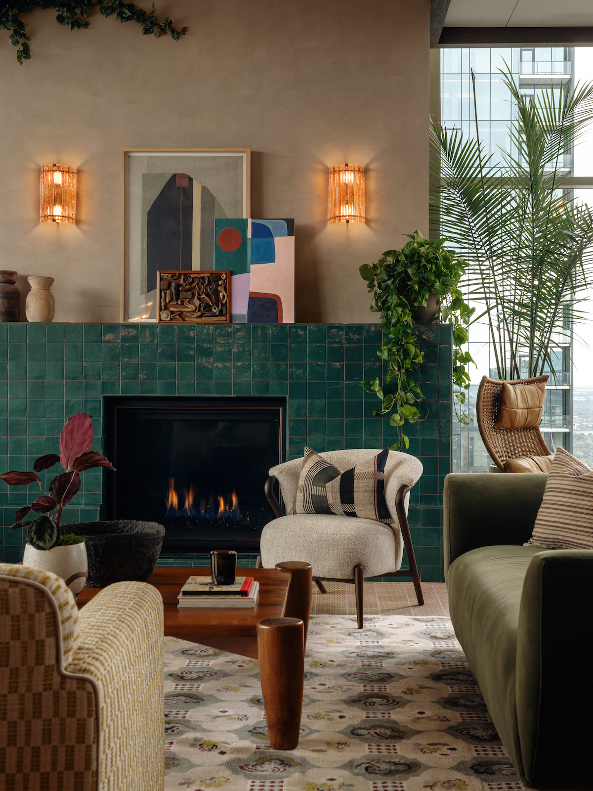 A stylish living room features a green tiled fireplace, abstract art, plants, cozy armchairs, and a green velvet sofa. Warm lighting and large Paseo windows create a cozy, modern atmosphere.