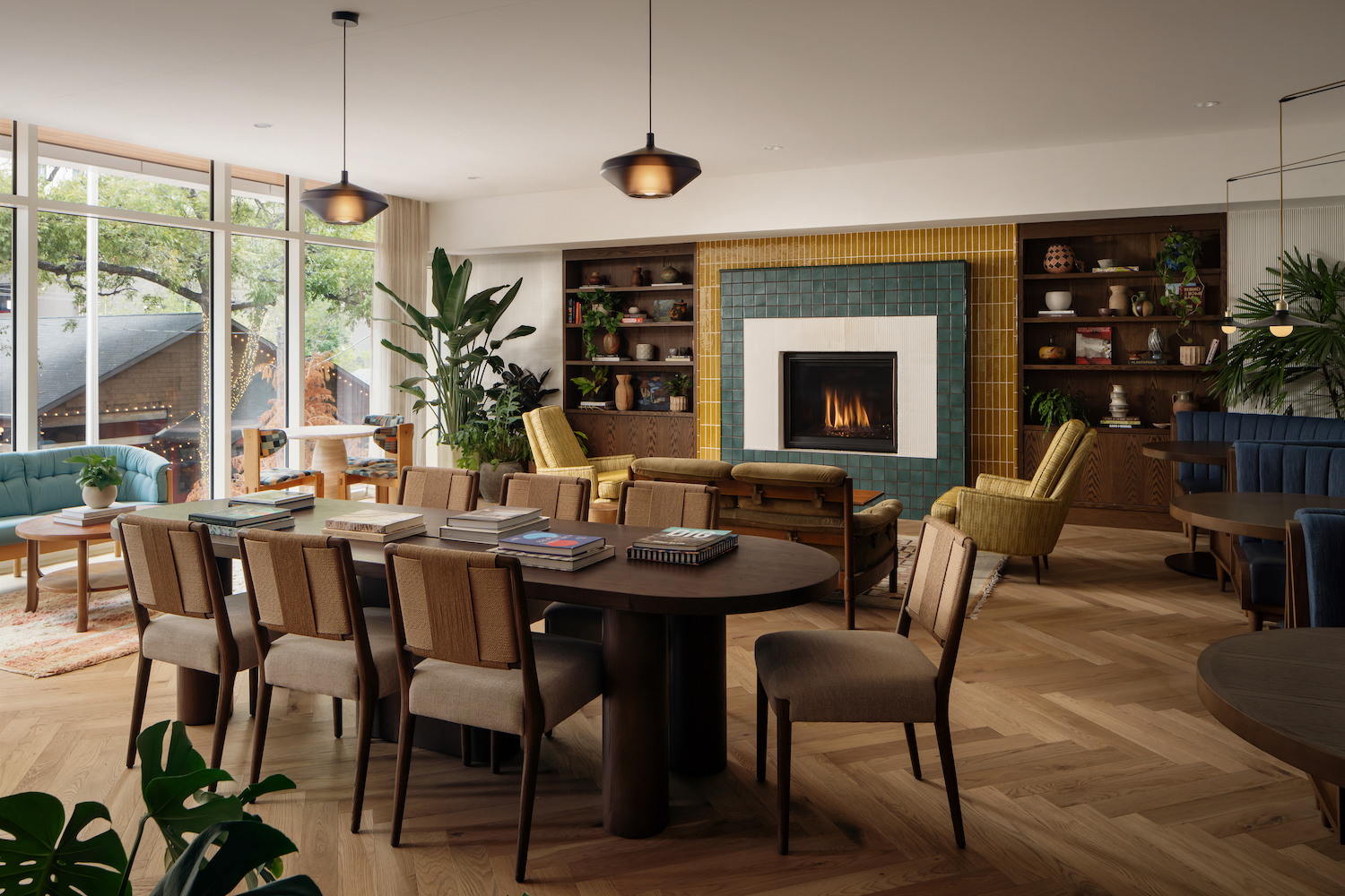 A stylish, modern living and dining area features a large wooden table with chairs, books, a green-tiled fireplace, cozy armchairs, built-in shelves, plants, and expansive windows offering a Paseo-like view of the trees outside.