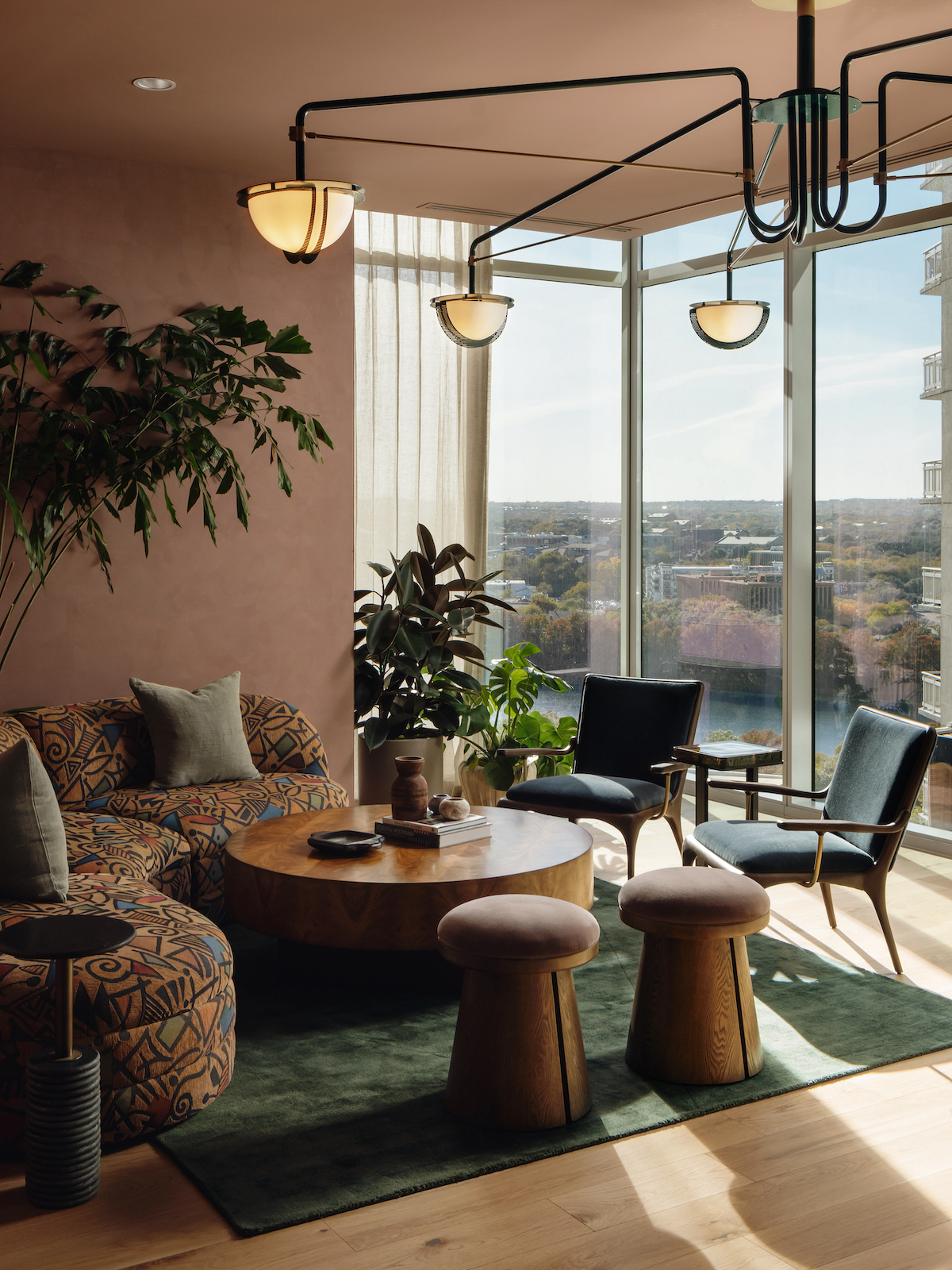 Modern living room with large windows overlooking the Paseo, featuring patterned sofas, velvet chairs, two round wooden stools, a low wooden coffee table, green rug, potted plants, and a black ceiling light. Sunlight streams in with cityscape views.