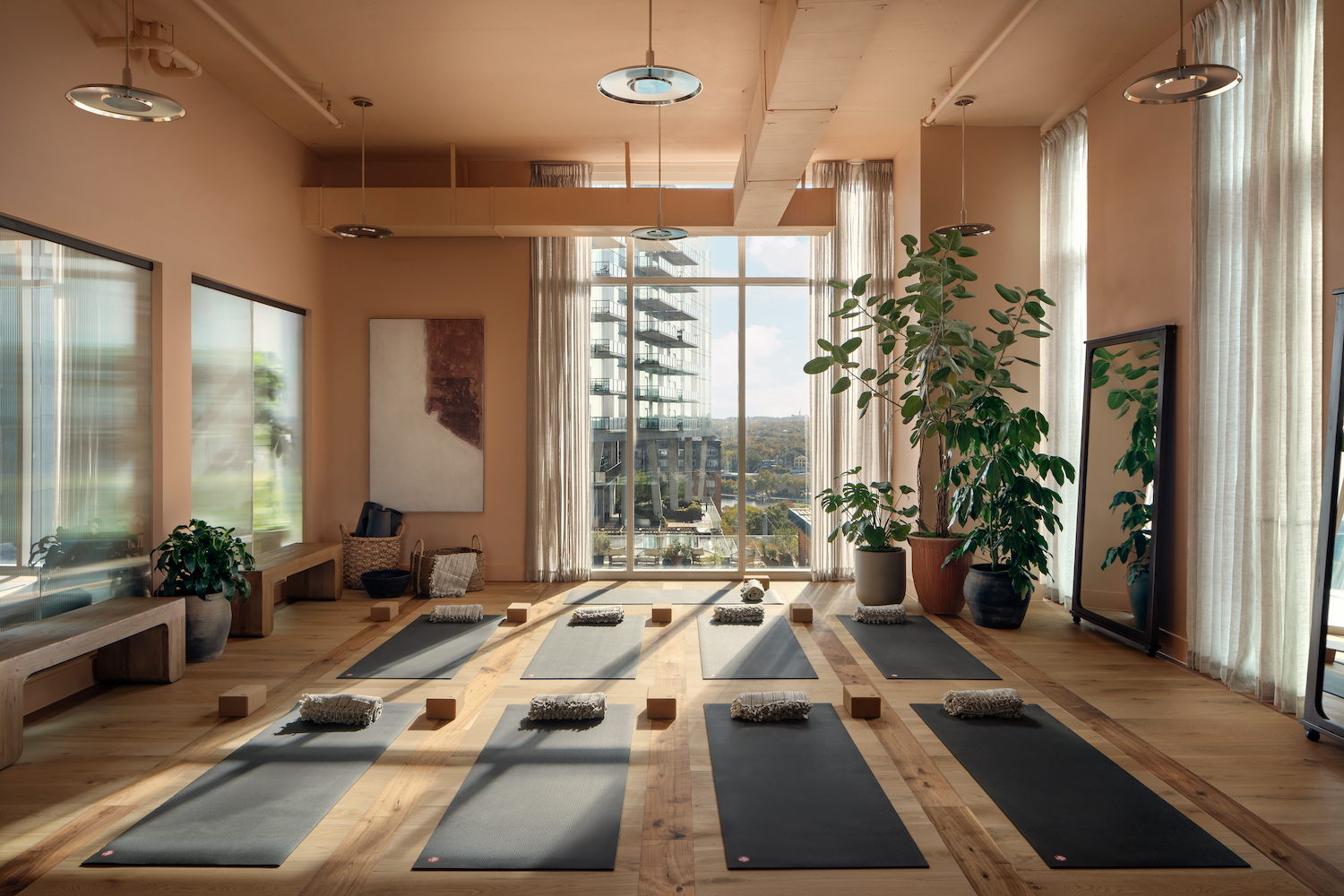 A bright, modern yoga studio on the Paseo with large windows, wooden floors, potted plants, and yoga mats neatly arranged with blocks and cushions. Natural light fills the room, and a cityscape is visible outside.