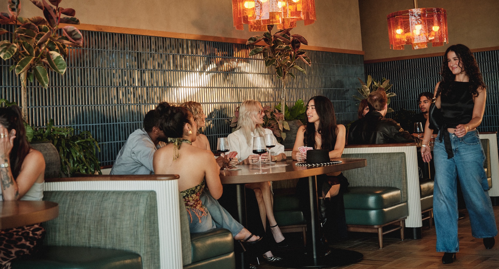 A group of people socialize and dine at a stylish restaurant with green booths, wooden floors, and warm orange pendant lights—echoing the chic interiors often found in luxury apartments Austin TX offers. One woman walks past as others converse at tables.