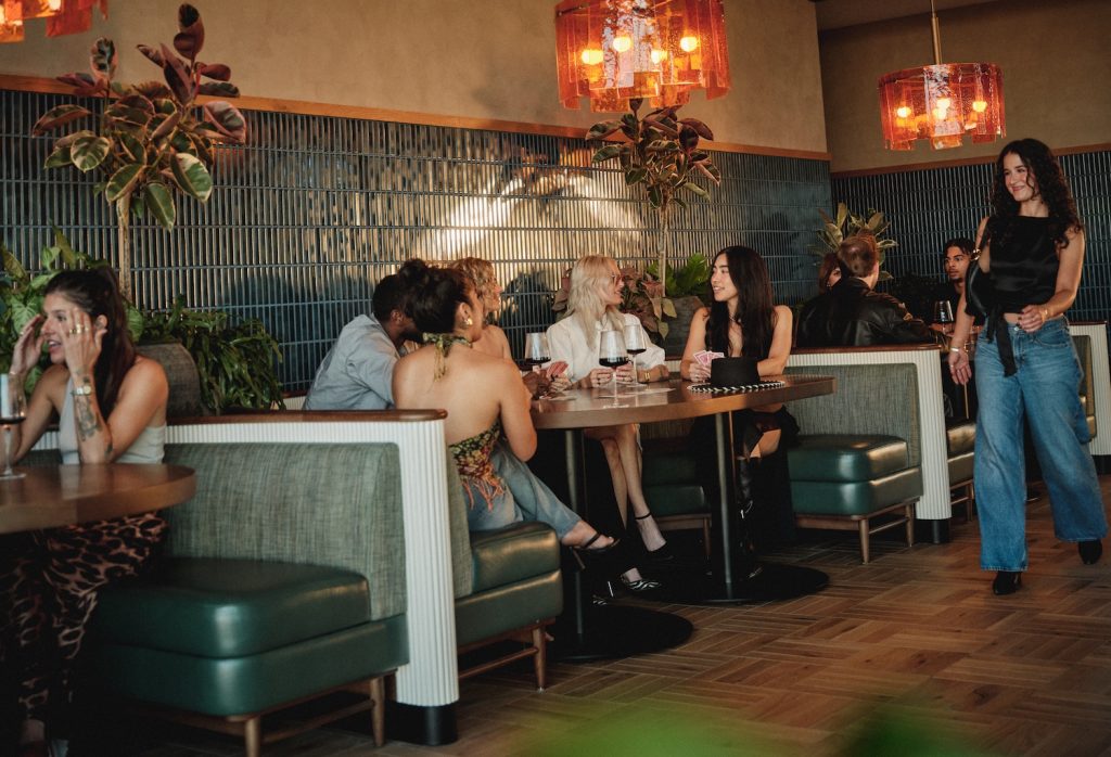 A group of people socialize and dine at a stylish restaurant with green booths, wooden floors, and warm orange pendant lights—echoing the chic interiors often found in luxury apartments Austin TX offers. One woman walks past as others converse at tables.