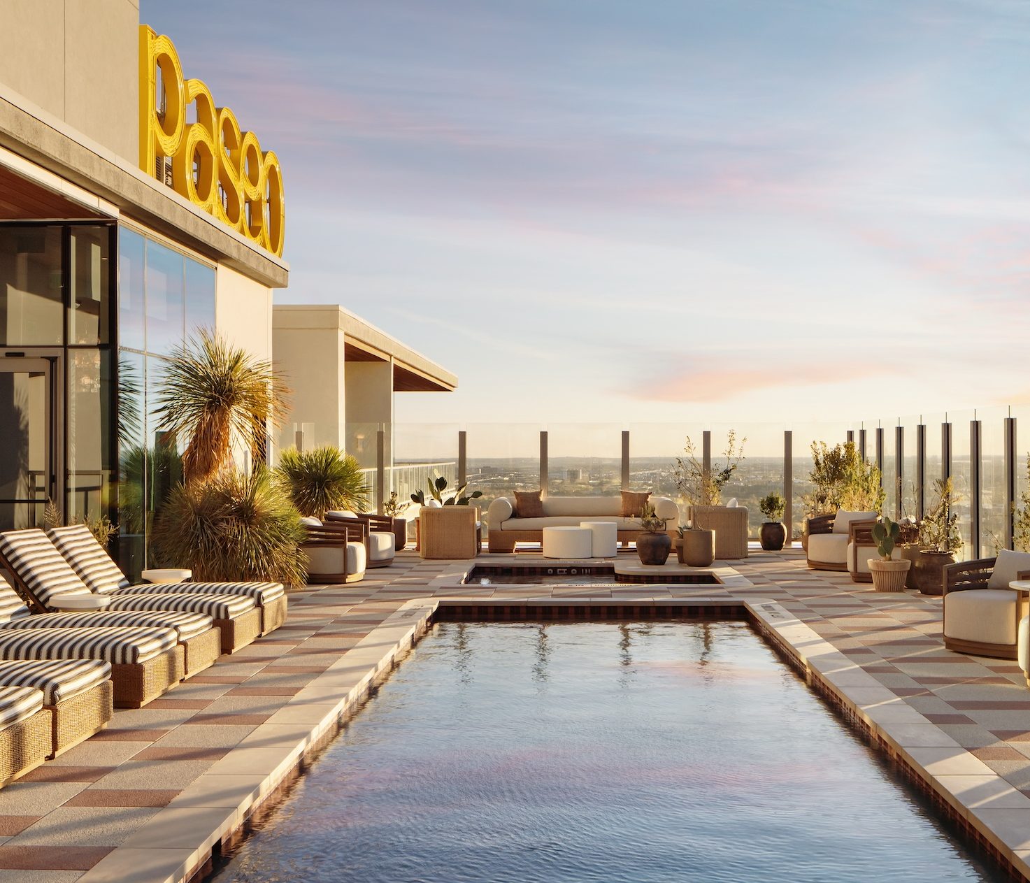A rooftop pool area designed for wellness, with striped lounge chairs, potted plants, and glass railing at sunset. Modern seating surrounds the pool, a large yellow sign is mounted on the building, and a cityscape sparkles in the background.