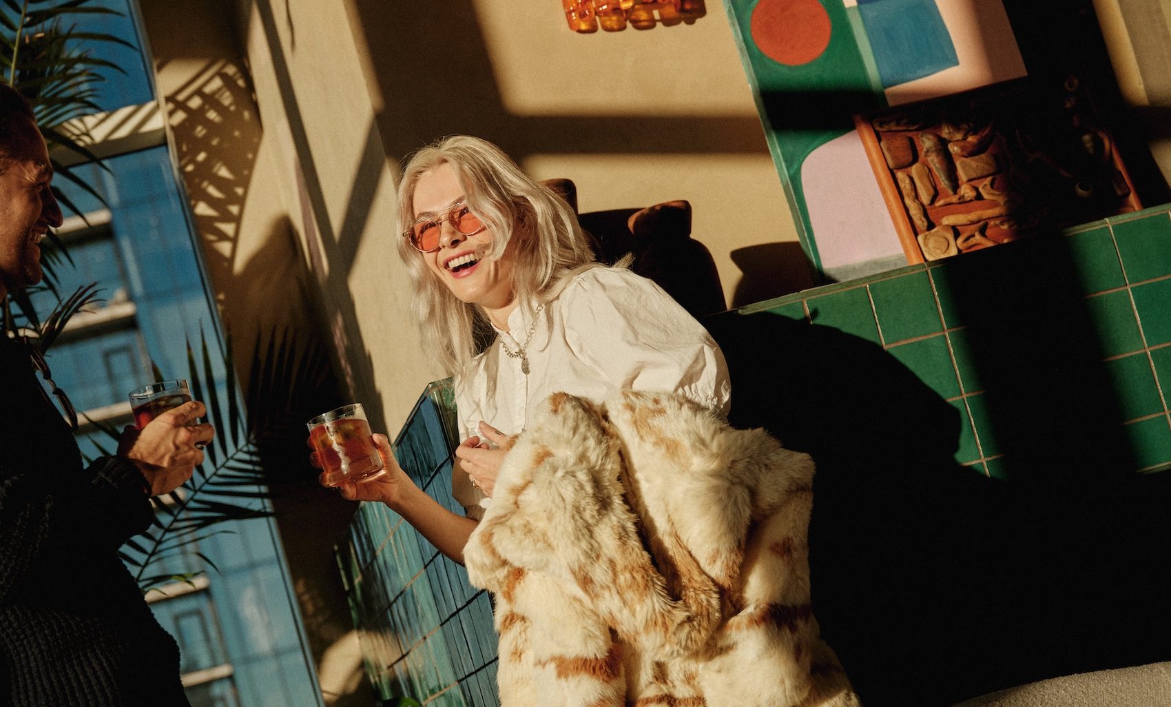 A woman with light hair, sunglasses, and a fur coat laughs while holding a drink indoors. Sunlight streams through the window, casting shadows on the green-tiled wall and decorative objects behind her.