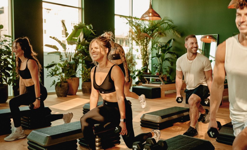 A group of people exercise indoors, smiling and performing lunges with dumbbells on step platforms in a bright, plant-filled gym that promotes wellness.