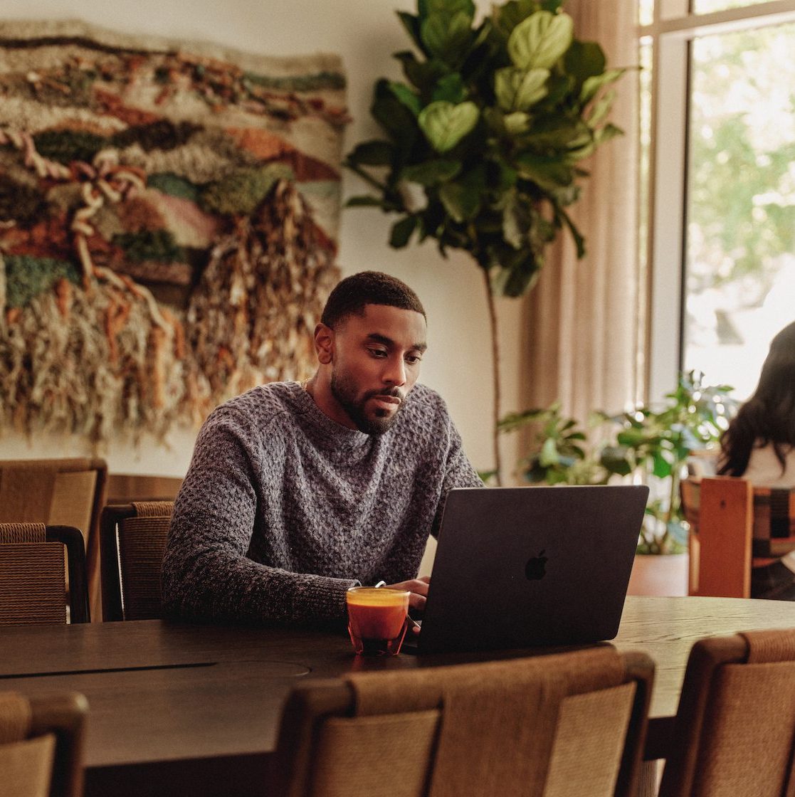 A man sits alone at a large table in a cozy, well-lit room, working on a laptop with a drink nearby. The stylish space, featuring a textile wall hanging and tall plant, reflects the charm of apartments in downtown Austin.