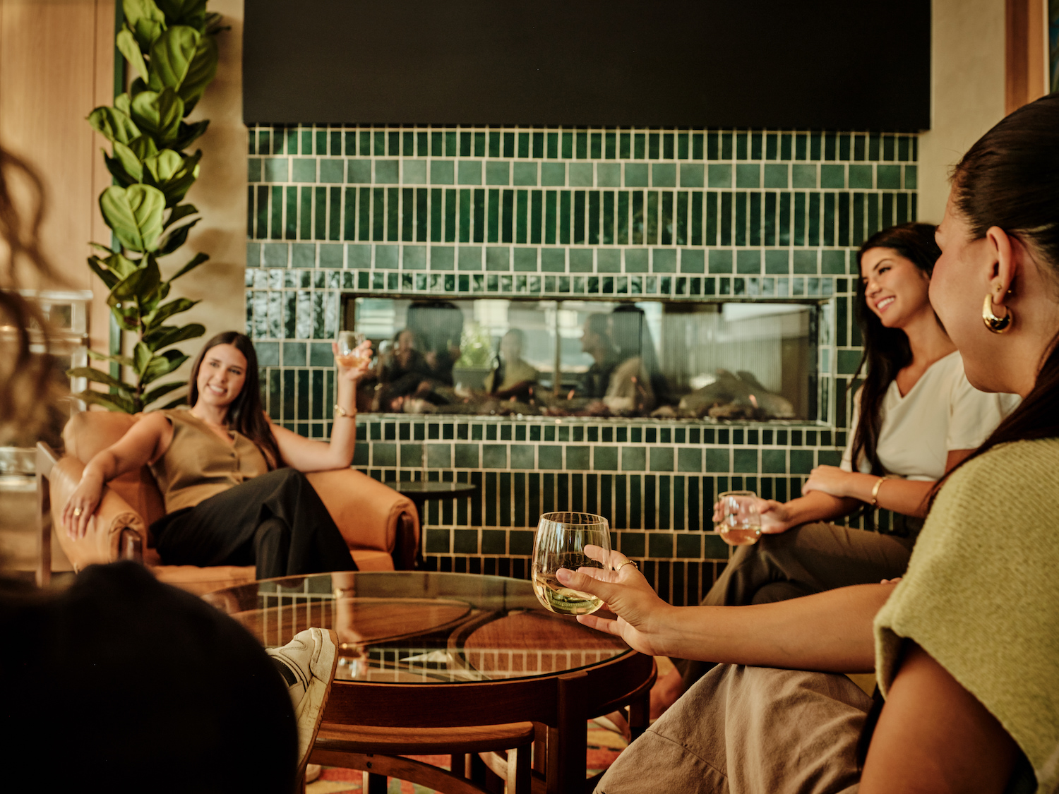 Four women sit and chat around a coffee table, each holding a drink, in a cozy room with a green-tiled fireplace—echoing the inviting ambiance of luxury apartments Austin TX—with lush plants creating a warm and relaxed atmosphere.