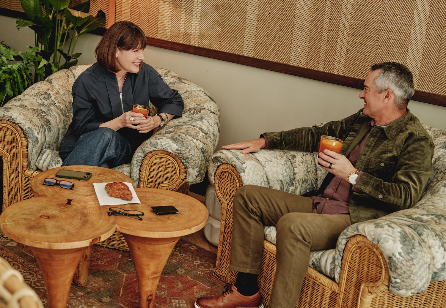 Two people sit in cozy wicker armchairs, smiling and holding drinks. A small wooden table with a pastry and their phones separates them. Warm lighting and plants create a relaxed, inviting vibe—just another day at luxury apartments Austin TX.