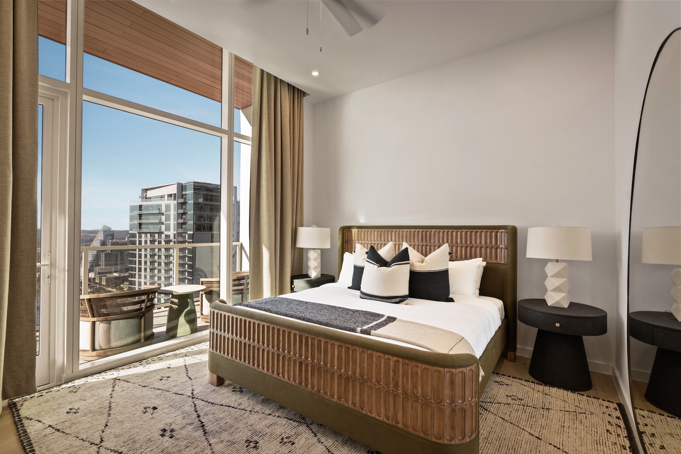 A modern Paseo Penthouse bedroom features a large bed, two bedside tables with lamps, a patterned rug, floor mirror, and floor-to-ceiling windows overlooking city buildings and a furnished balcony.