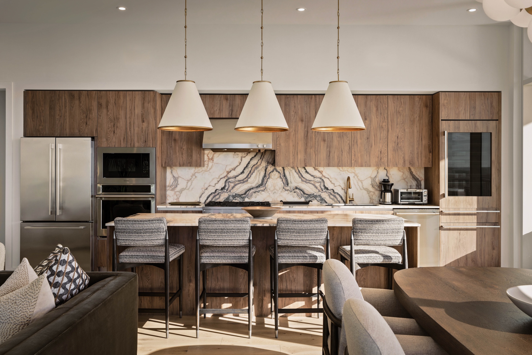 Modern kitchen with wood cabinets, marble backsplash, three pendant lights, stainless steel appliances, and a kitchen island with four gray barstools—an inviting space in the Paseo Penthouse, with the dining table and sofa visible in the foreground.