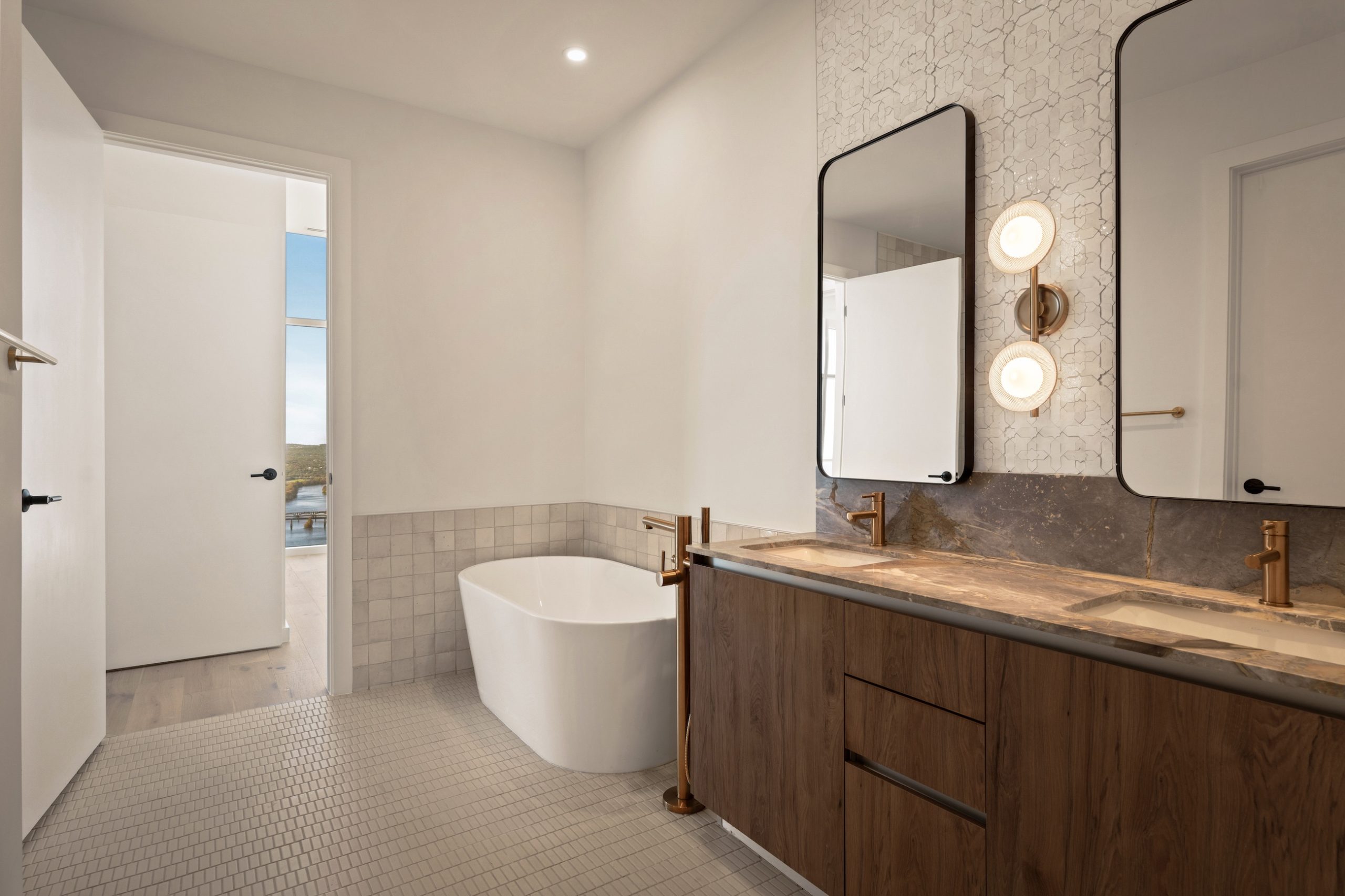 Modern paseo penthouse bathroom with a freestanding white tub, double vanity with wood cabinets, two rectangular mirrors, gold fixtures, and wall-mounted lights; door open to a view outside.