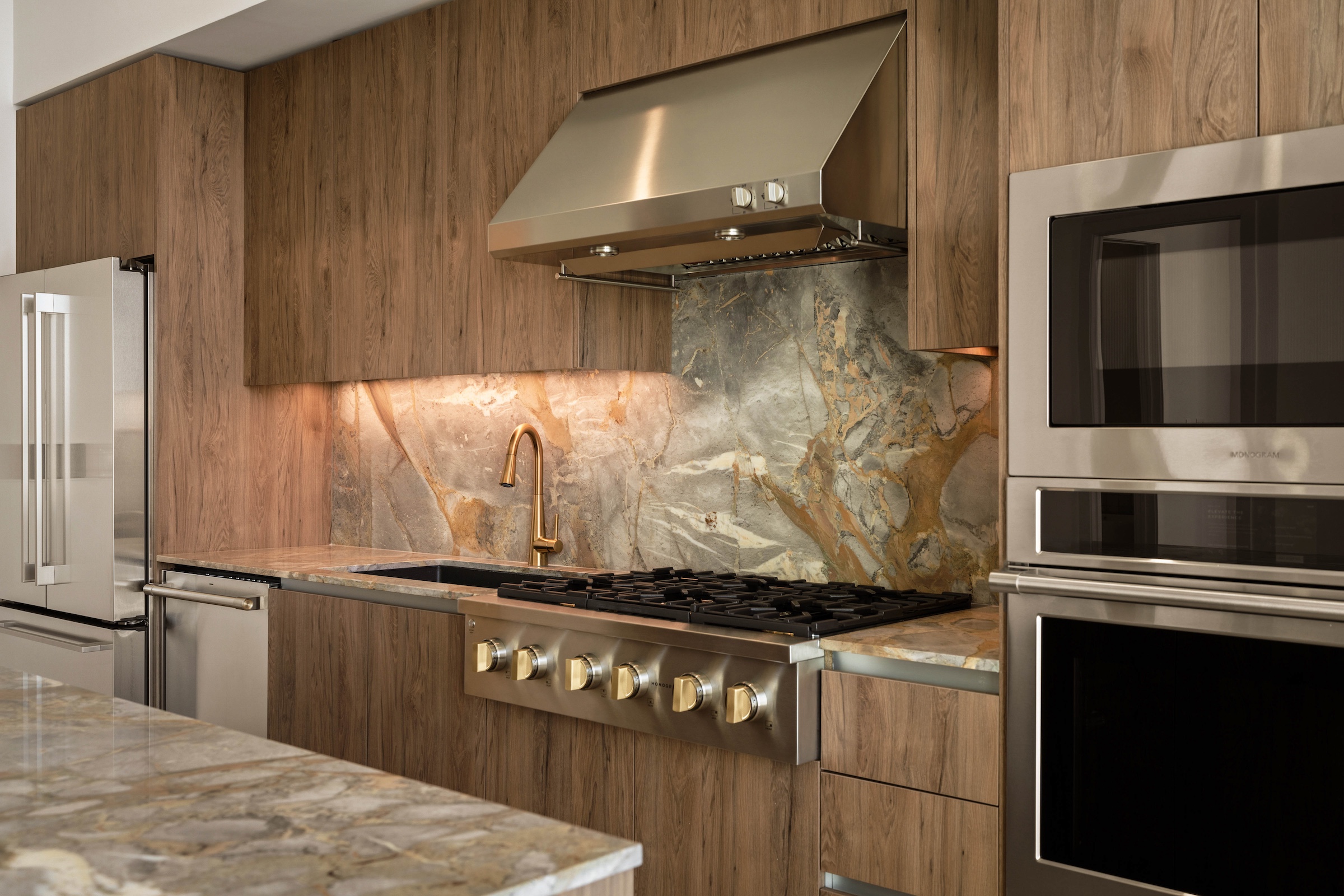 Modern paseo penthouse kitchen with wood-paneled cabinets, stainless steel appliances, a large gas stove, sleek range hood, and stone backsplash and countertop in earthy tones. Warm lighting highlights the backsplash area.