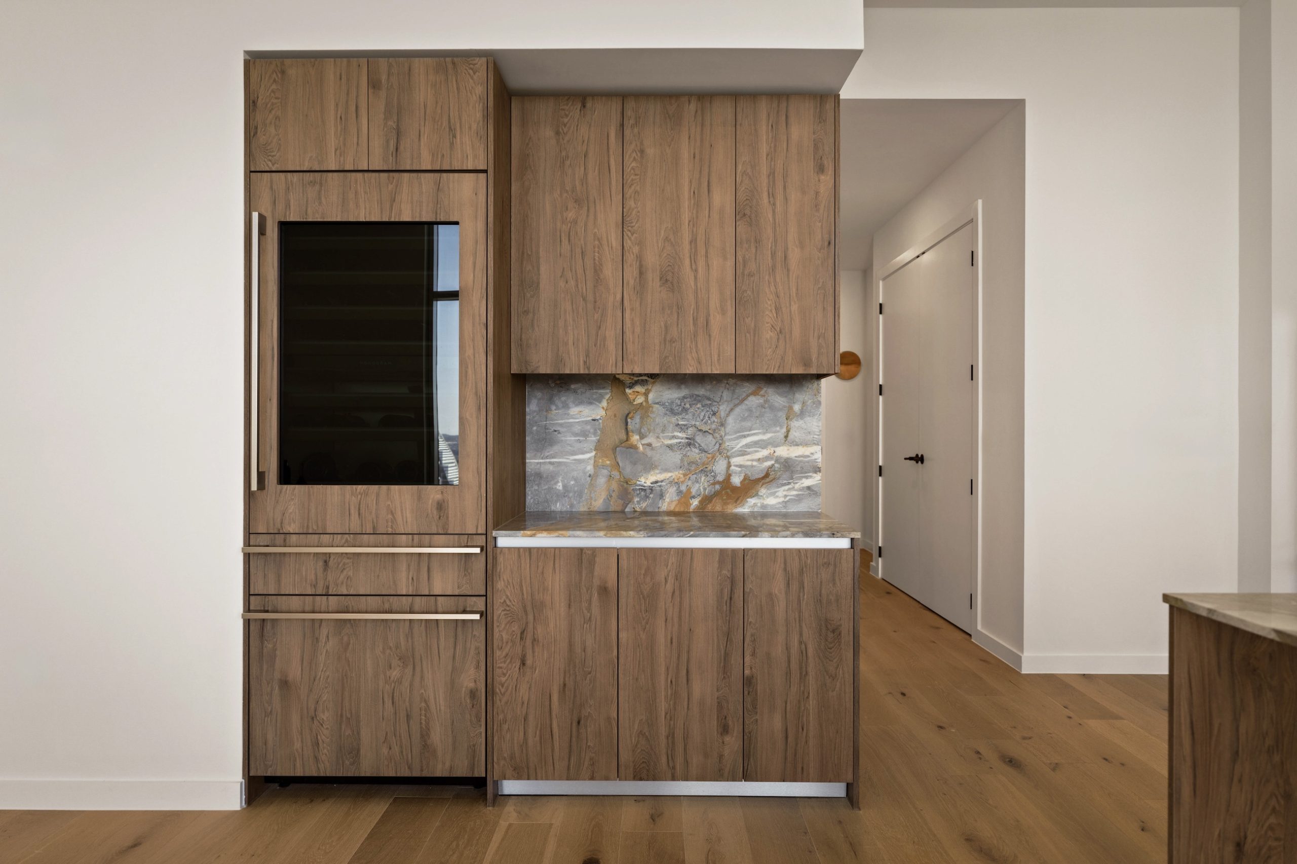 A modern kitchen in the paseo penthouse features wood cabinets, a built-in dark glass refrigerator, a marble backsplash with brown and gray tones, and light wood flooring. White walls and a hallway with closed doors are visible in the background.