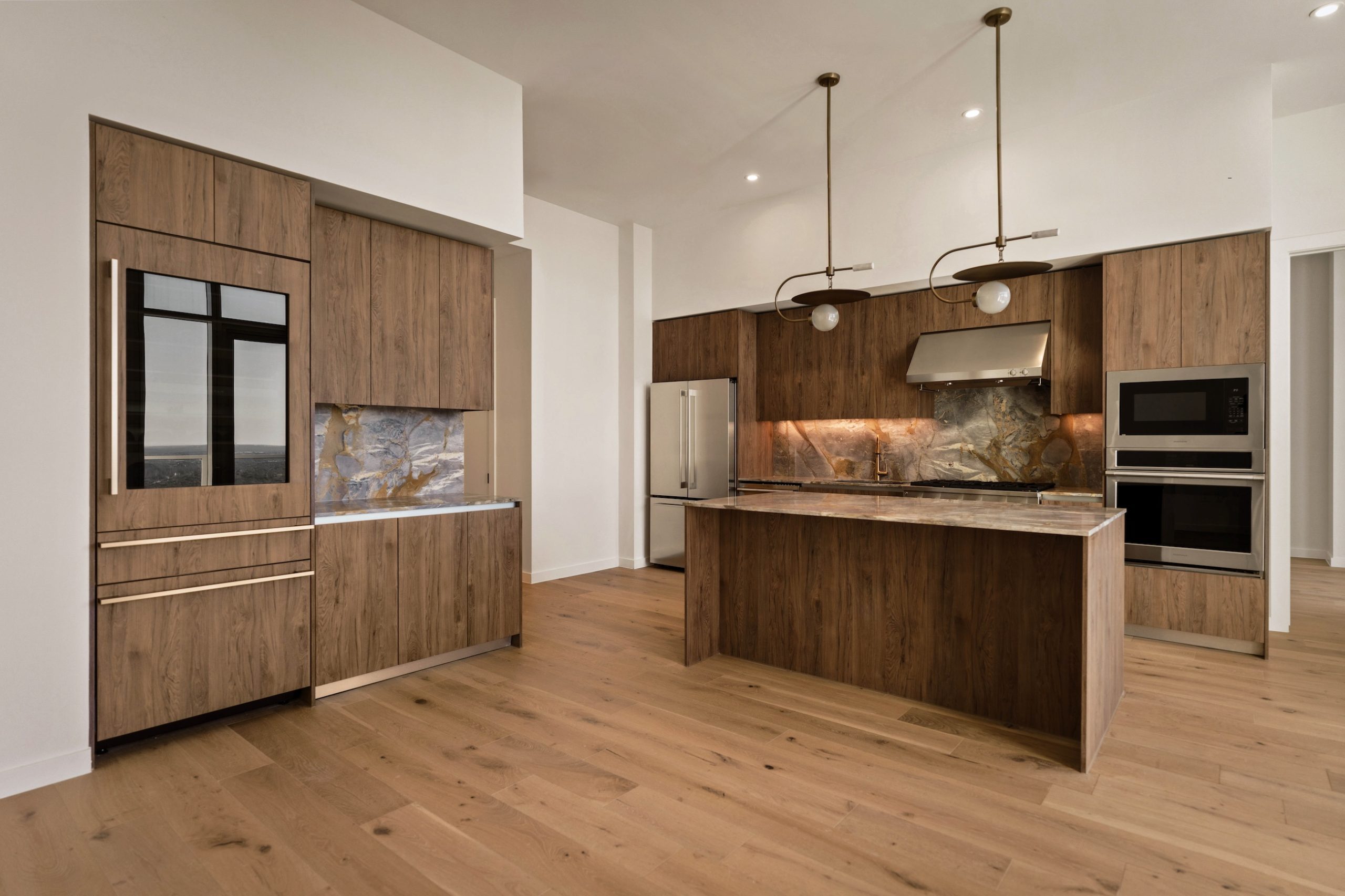 Modern kitchen in the Paseo Penthouse featuring wood cabinets, marble backsplash and countertops, an island with pendant lights, built-in oven and microwave, stainless steel fridge, and light wood flooring. The open space is bright and inviting.