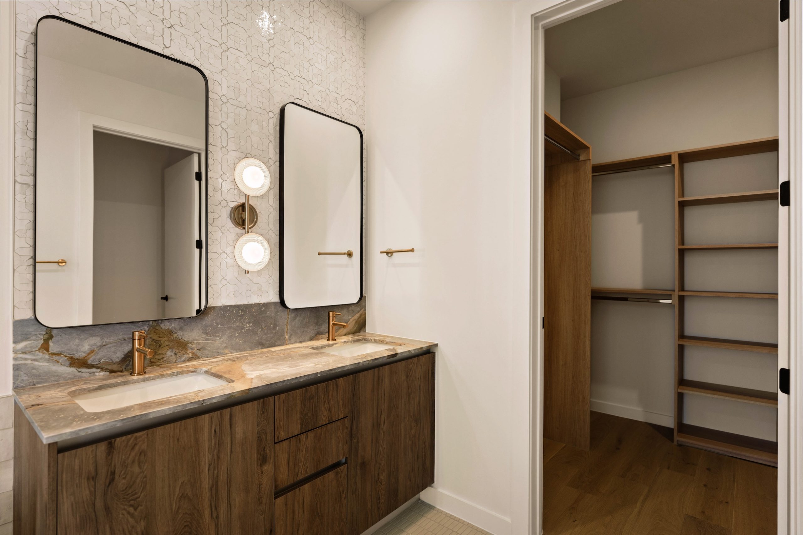 Modern bathroom in the Paseo Penthouse with a double sink vanity, wood cabinets, marble countertop, two rectangular mirrors, gold faucets, decorative wall lighting, and an adjacent walk-in closet with built-in wooden shelves and rods.