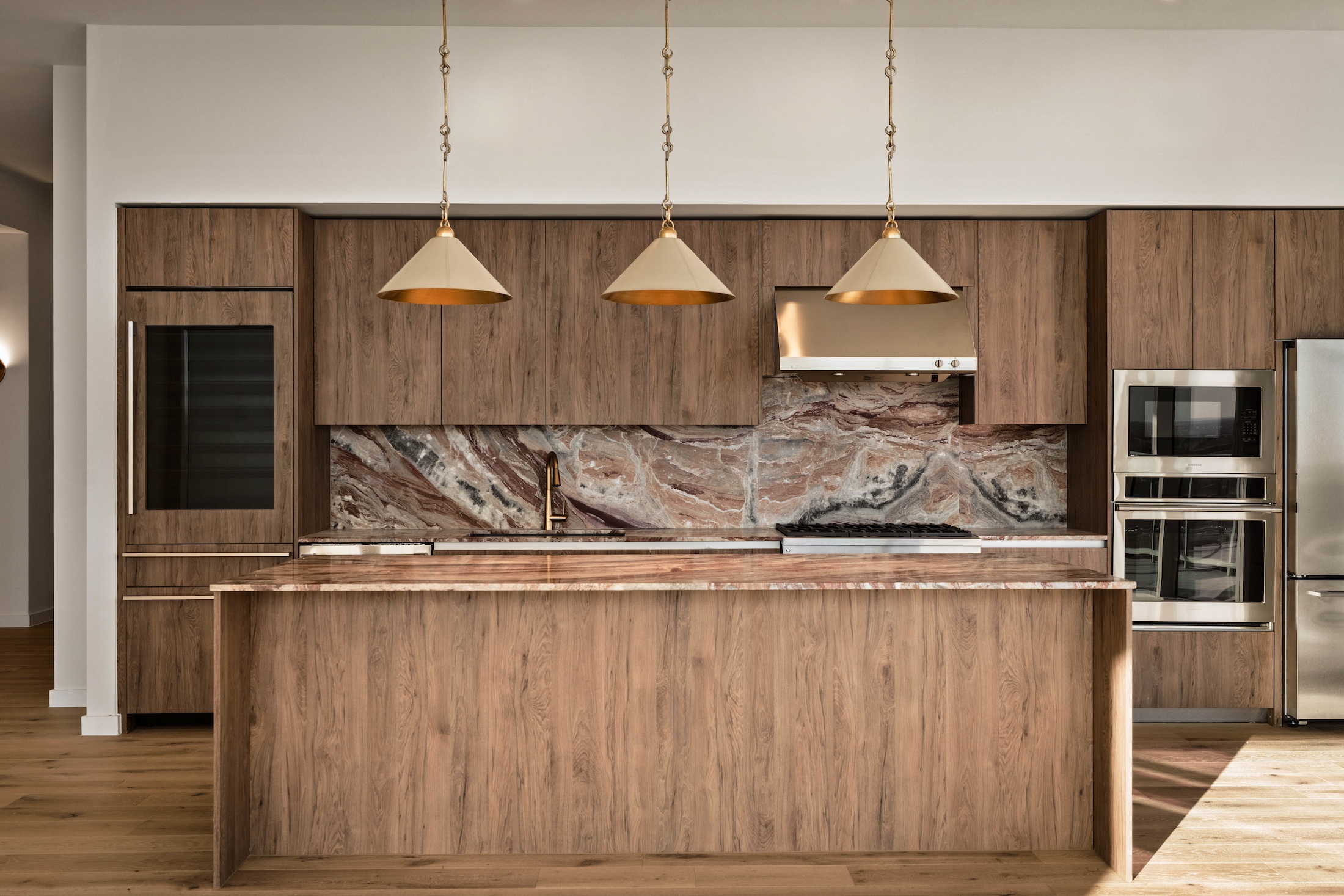Modern kitchen with wood cabinets, a marble backsplash and countertop, three gold pendant lights above the island, stainless steel appliances, and a built-in oven and fridge. The paseo penthouse features a clean, minimalist design.