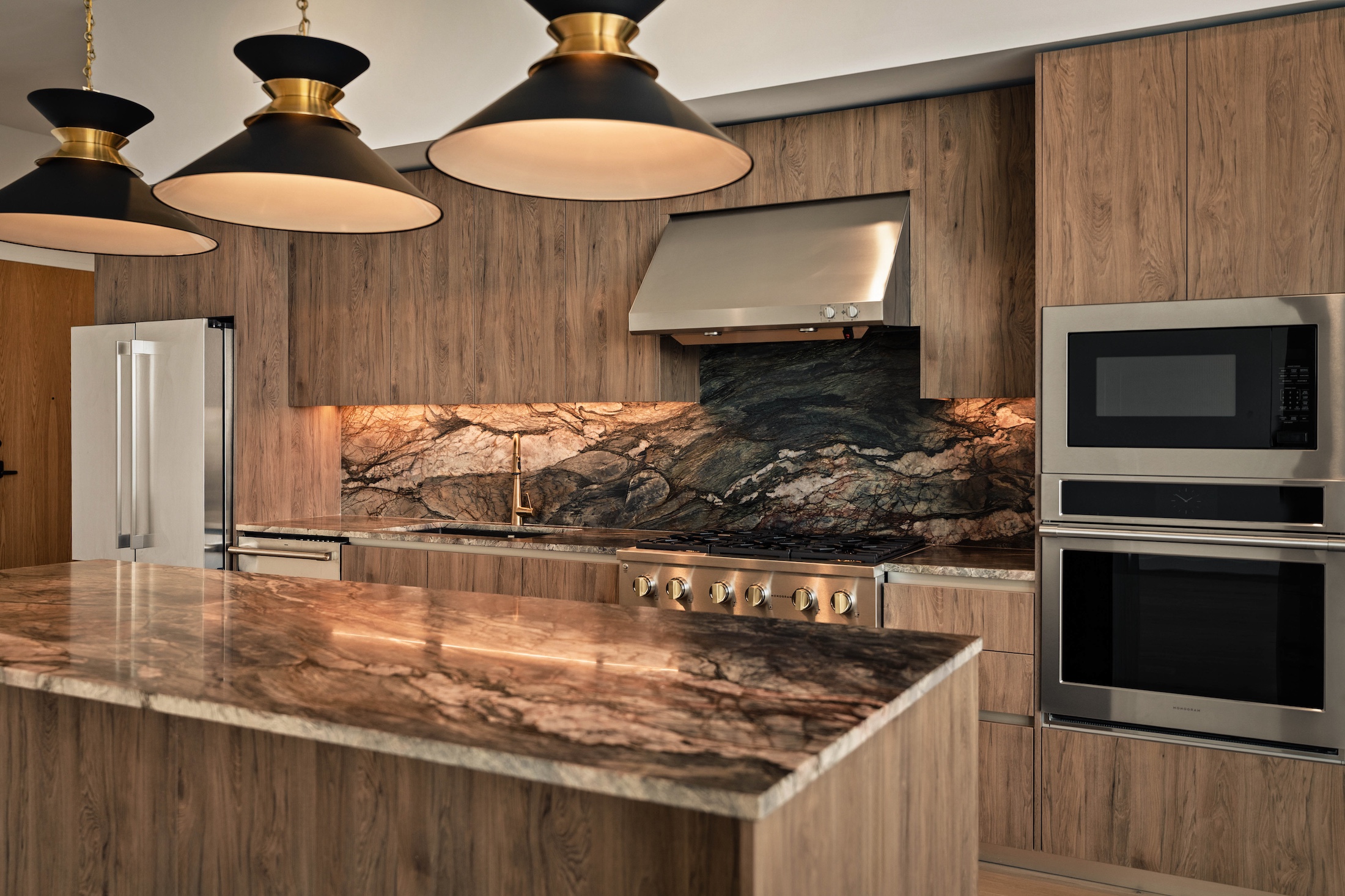 Modern kitchen in the Paseo Penthouse with wood cabinetry, marble countertops and backsplash, a stainless steel stove, double oven, microwave, and three large black and gold pendant lights over an island.