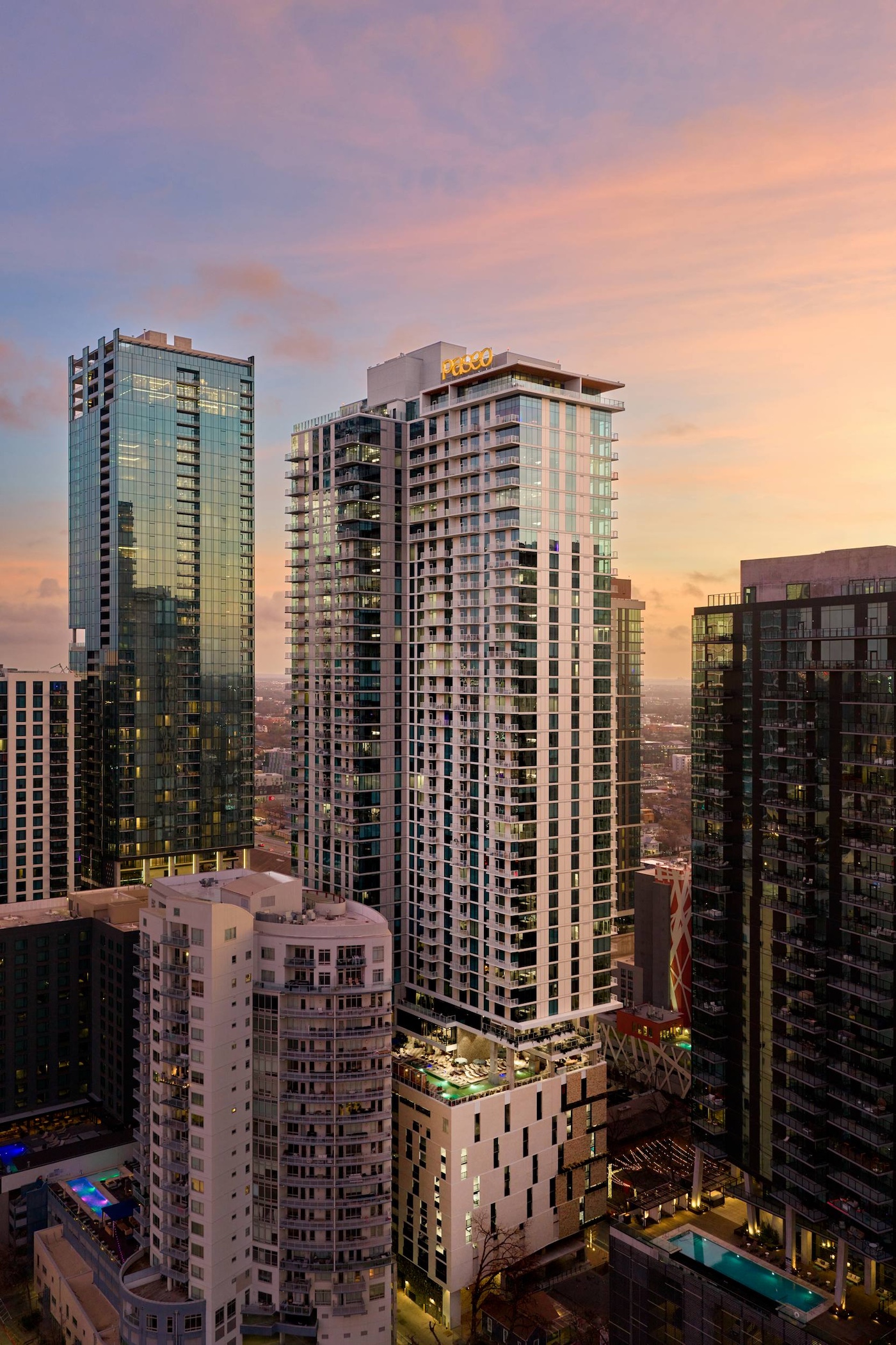 A cluster of modern high-rise buildings lines the city at sunset, with the tallest displaying “PLUGO” on top. Vibrant shades of orange, pink, and blue fill the sky above this bustling paseo.
