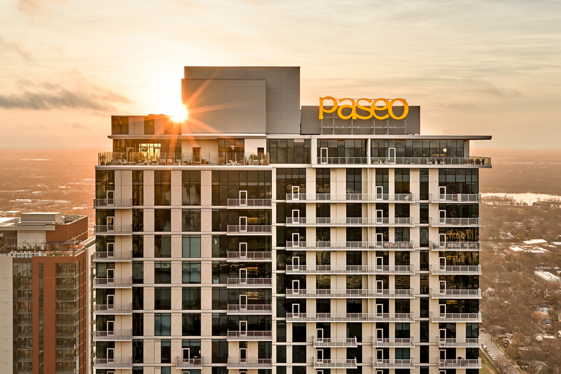 A modern high-rise building with “Paseo” in large yellow letters atop, shown at sunset as sunlight peeks around the rooftop, with cityscape and trees framing the sleek “Paseo” against the evening sky.