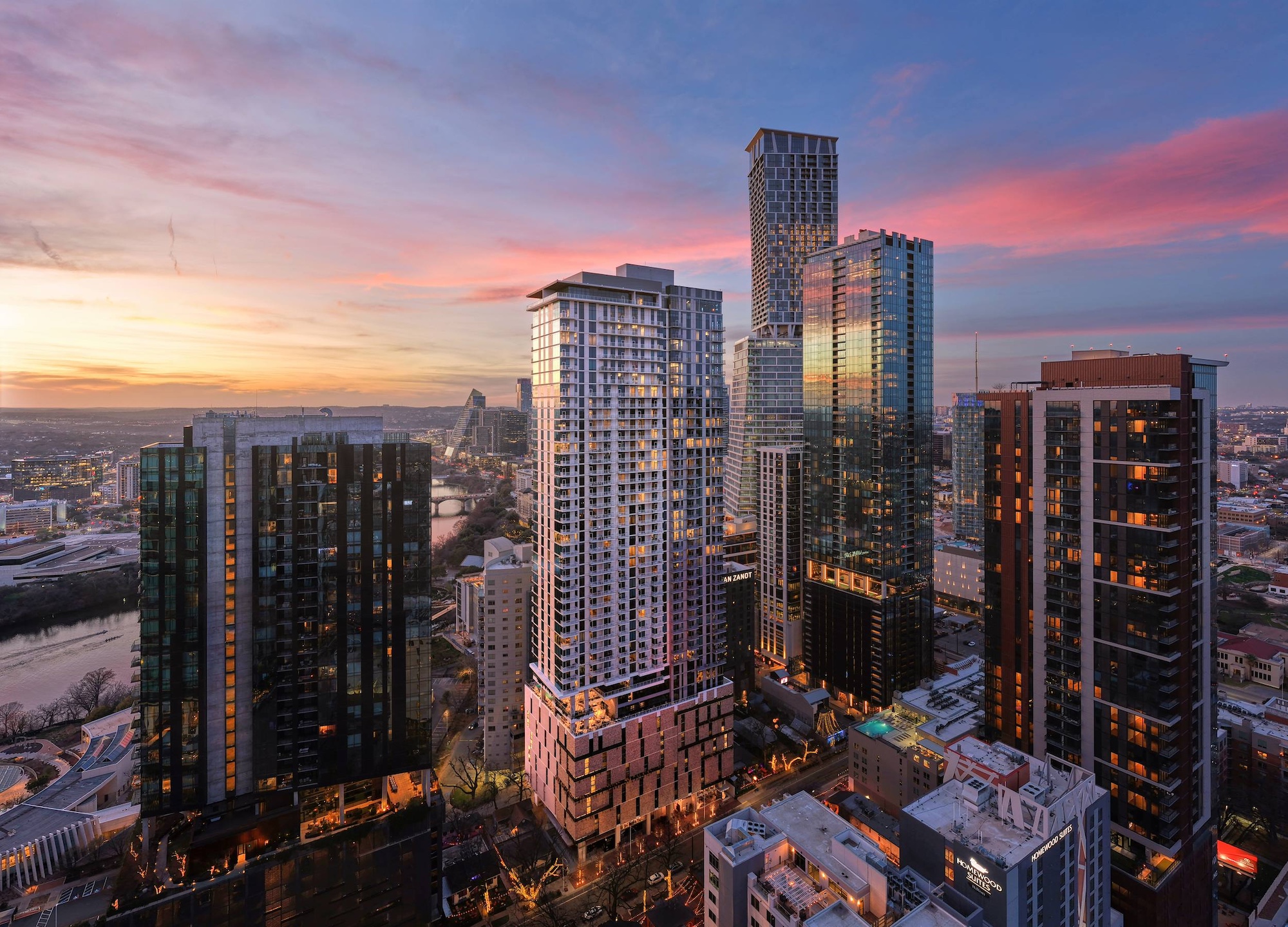 A cityscape at sunset with tall modern skyscrapers and glowing glass exteriors, set above a riverside paseo, as vibrant pink, orange, and blue hues fill the sky.