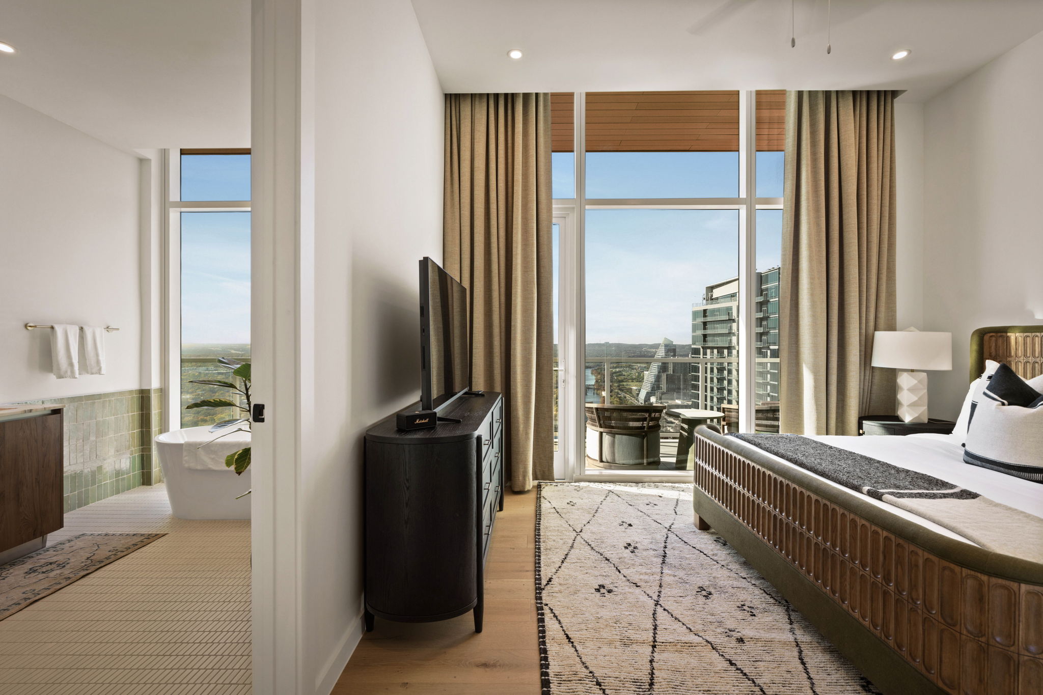 Modern paseo penthouse hotel room with a large bed, TV on a cabinet, and floor-to-ceiling windows leading to a balcony overlooking the city. An open bathroom with bathtub sits on the left, surrounded by neutral tones and natural light.