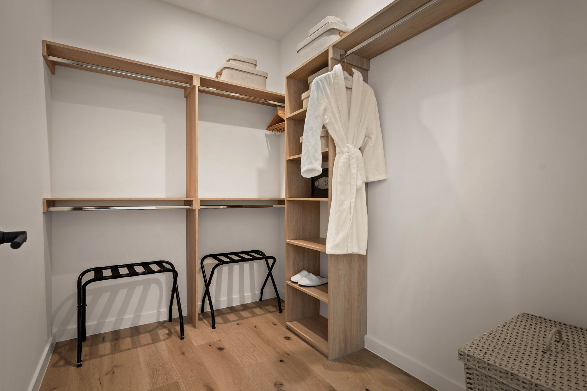 A modern walk-in closet in the Paseo Penthouse features wooden shelves and hanging rods, two folded towels on the top shelf, a white robe and slippers, two black luggage racks, and a woven laundry hamper on a wooden floor.