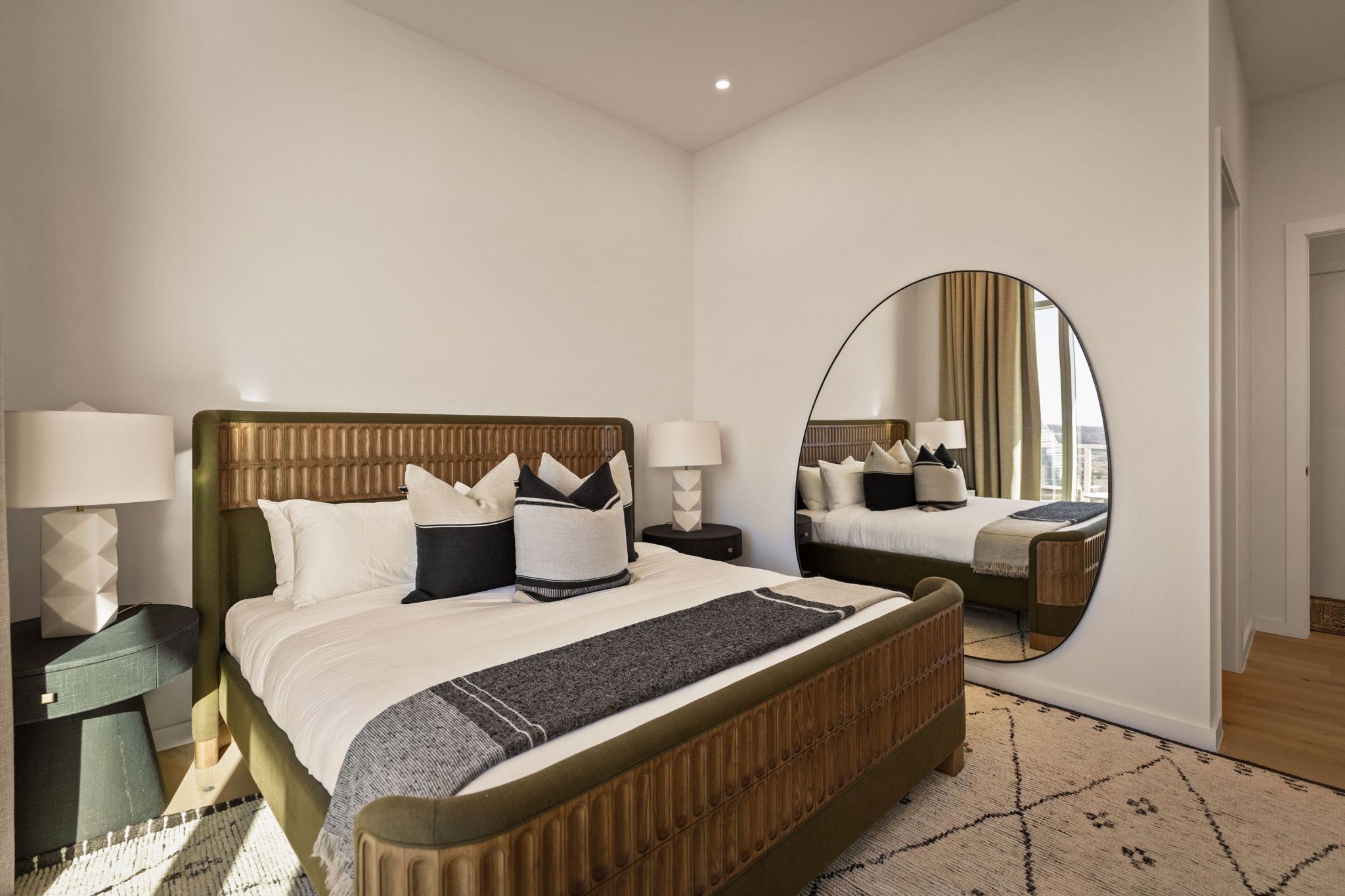 Modern paseo penthouse bedroom with a large bed, dark frame, neutral bedding, black accent pillows, two bedside tables with white lamps, a round mirror reflecting the room, and a patterned rug on light wood floors.