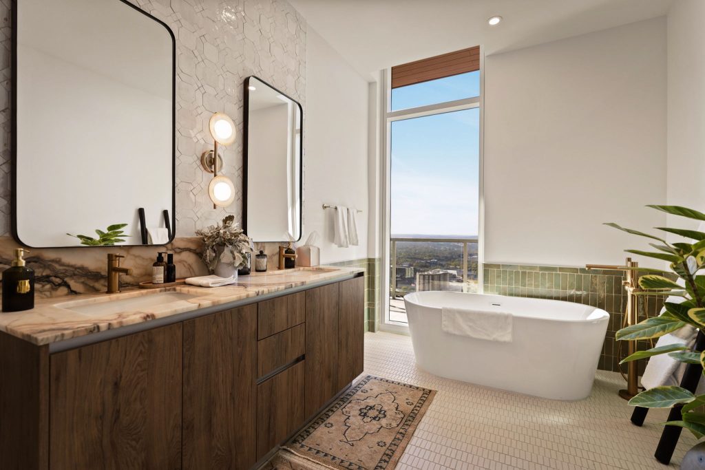 Modern bathroom in the Paseo Penthouse with double sinks, large mirrors, a freestanding tub near tall windows offering a city view, and green plants adding a touch of nature. Marble countertops and wood accents create a warm, elegant atmosphere.