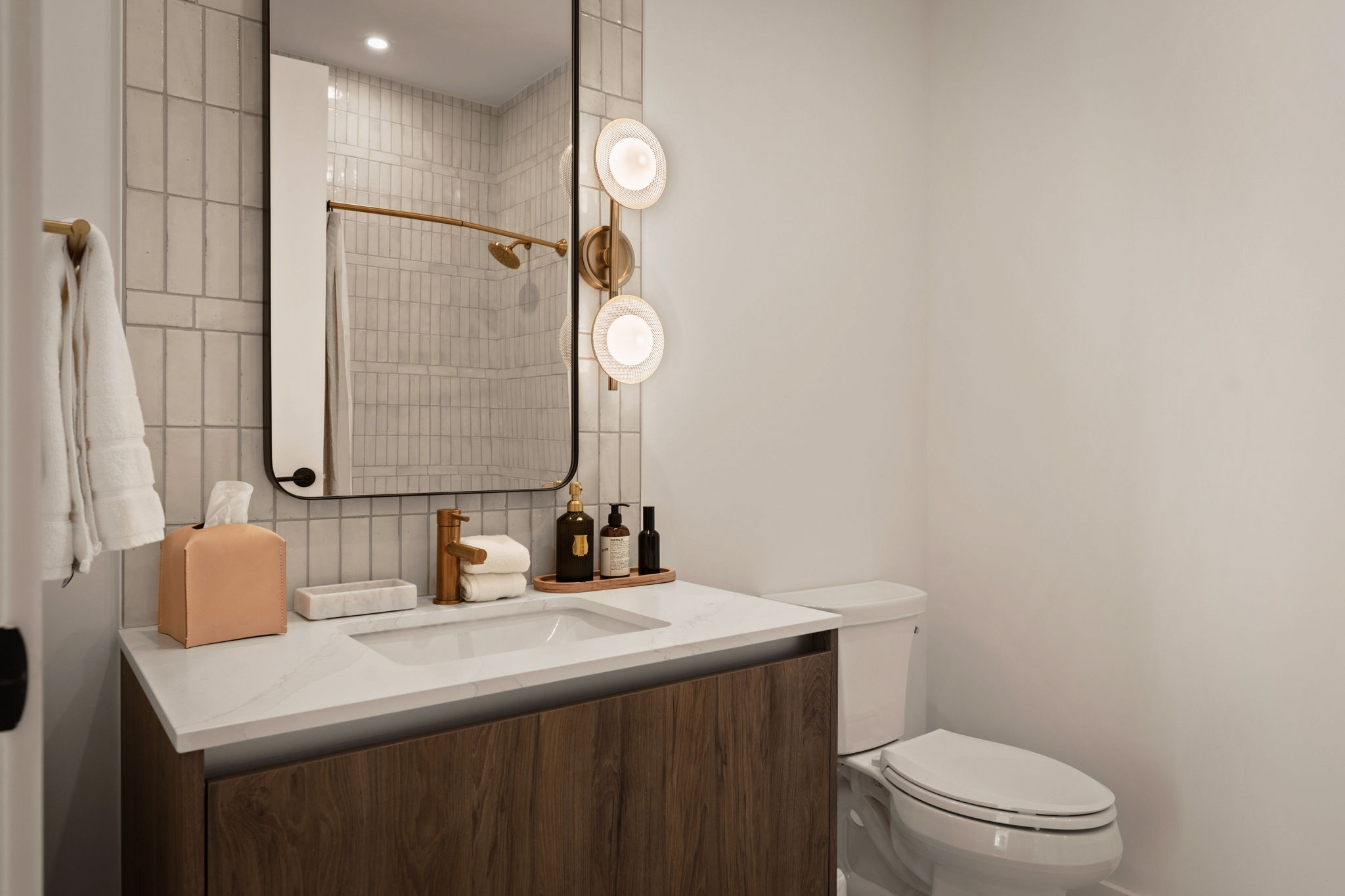 Modern bathroom in the Paseo Penthouse featuring a wood vanity, white countertop, mounted rectangular mirror, round wall lights, white toilet, and a shower with gold fixtures and white tile walls. Neutral tones and minimalist decor complete the space.
