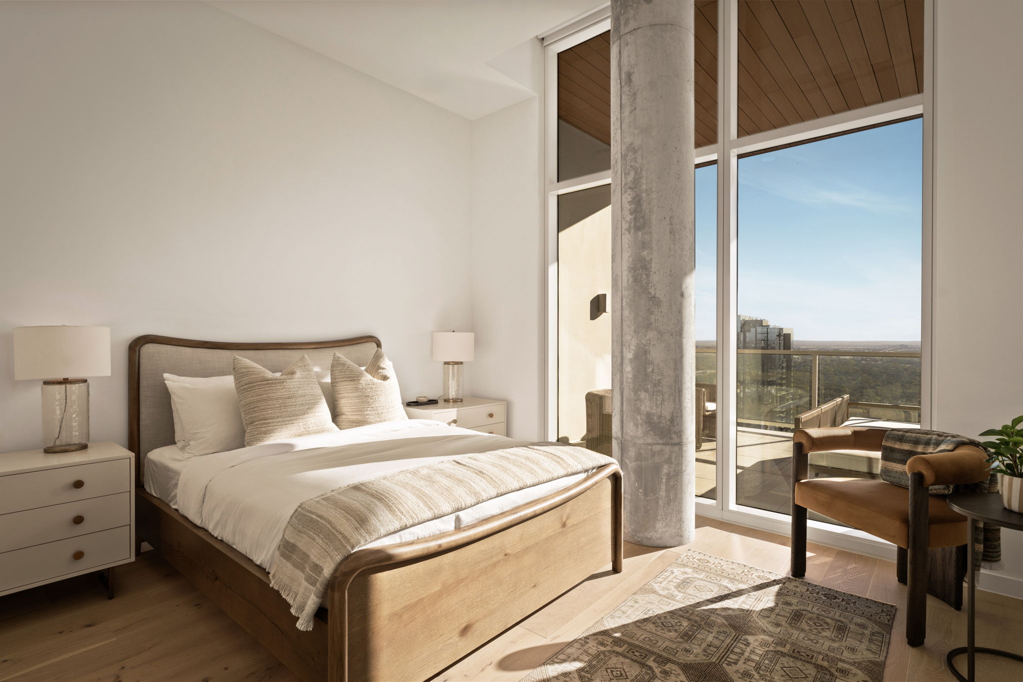 A modern bedroom in the Paseo Penthouse features a wooden bed, white bedding, two bedside tables with lamps, a patterned rug, a chair, and large windows opening to a balcony with a scenic view.