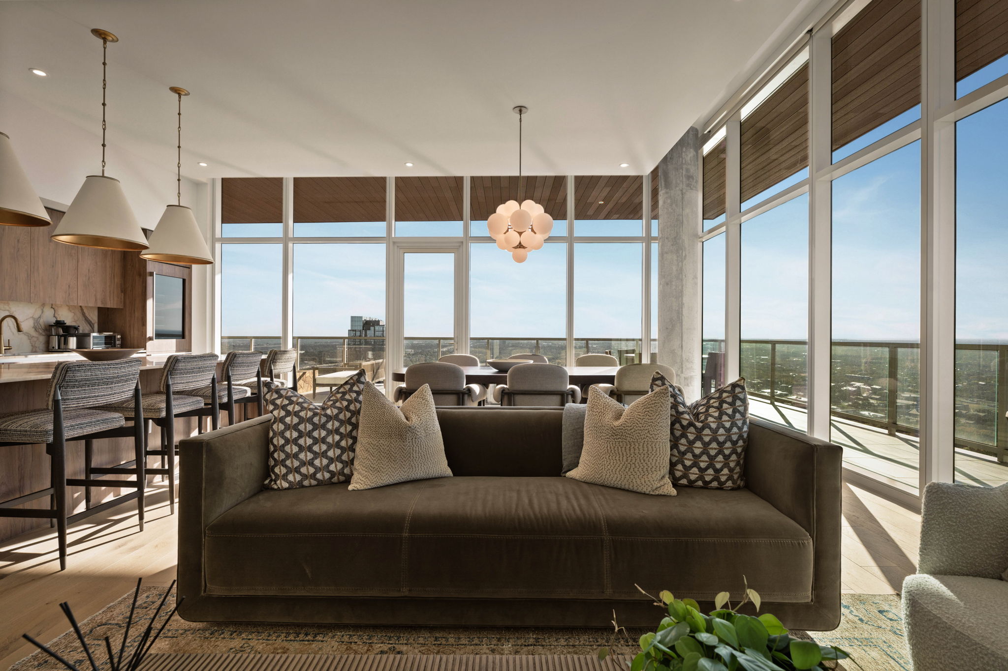 A modern, sunlit paseo penthouse living room with floor-to-ceiling windows, a dark sofa with patterned pillows, a dining table, and a kitchen area with bar stools. Light wood floors and city views complete the stylish space.