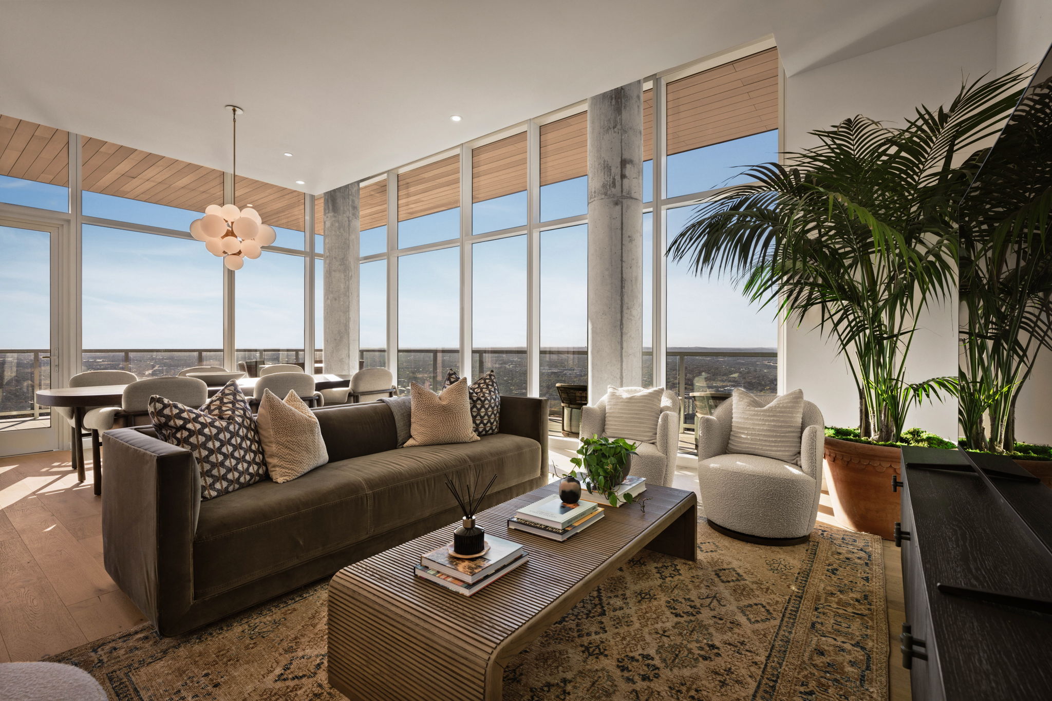 A modern, sunlit paseo penthouse living room with floor-to-ceiling windows, a gray sofa, patterned cushions, a coffee table with decor, two armchairs, large green plant, dining table, and city views in the background.