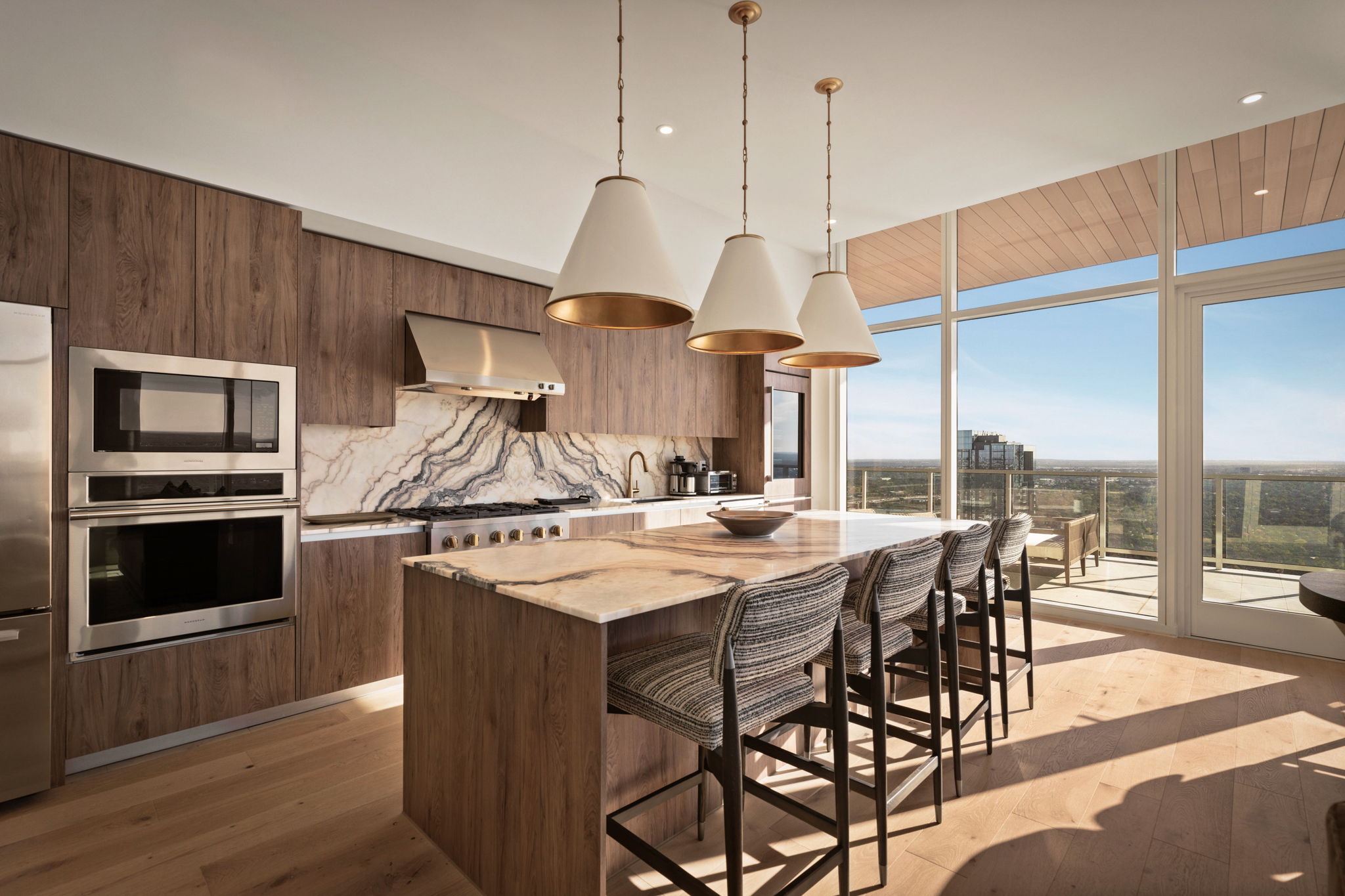 Modern kitchen in the Paseo Penthouse with wood cabinets, marble island and backsplash, stainless steel appliances, three pendant lights, and four woven barstools. Large windows offer a scenic view and fill the space with natural light.
