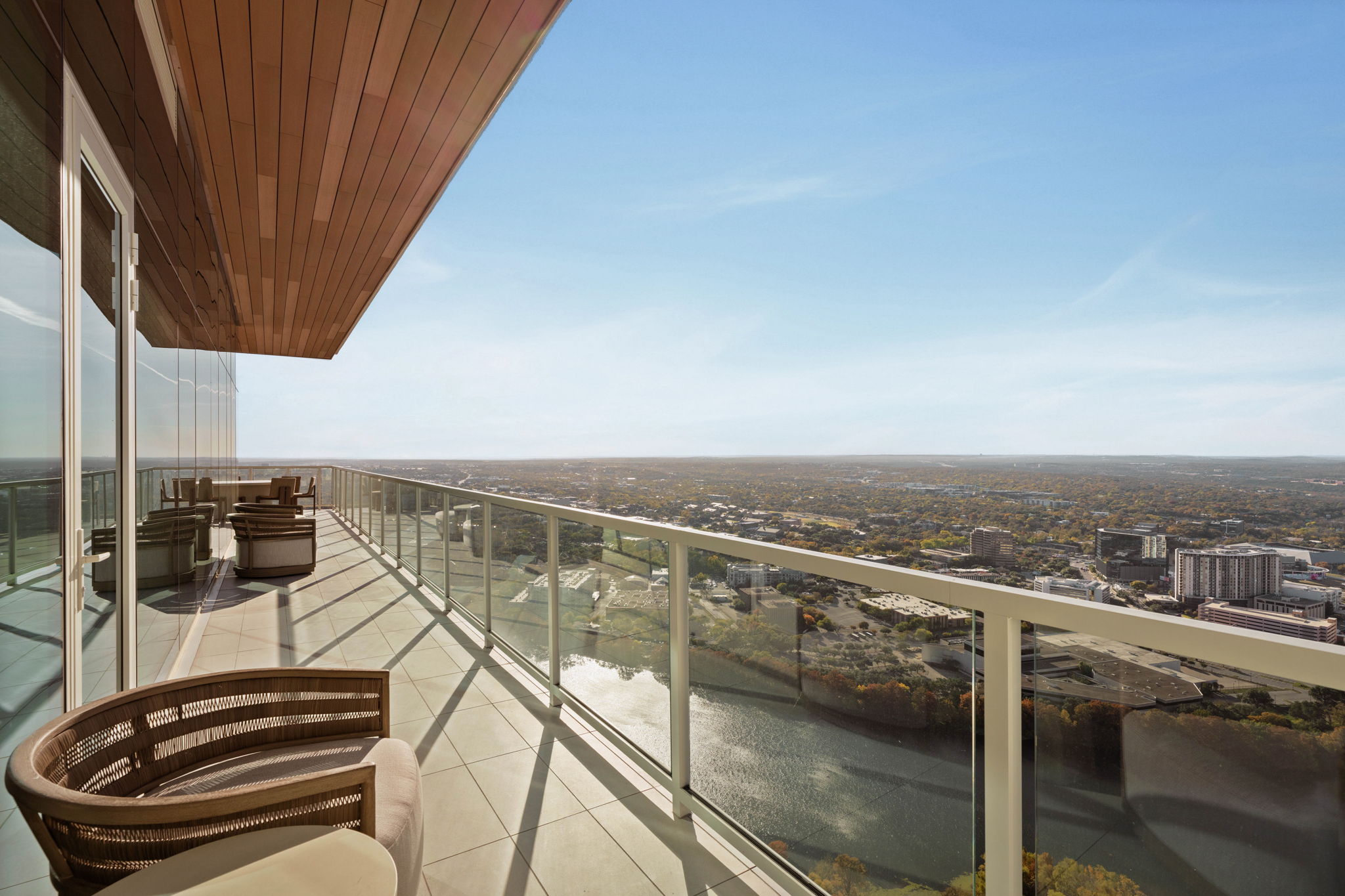 A spacious balcony in the Paseo Penthouse, with rattan chairs and glass railing, overlooks a cityscape, river, and clear blue sky—featuring outdoor seating and modern architectural details.