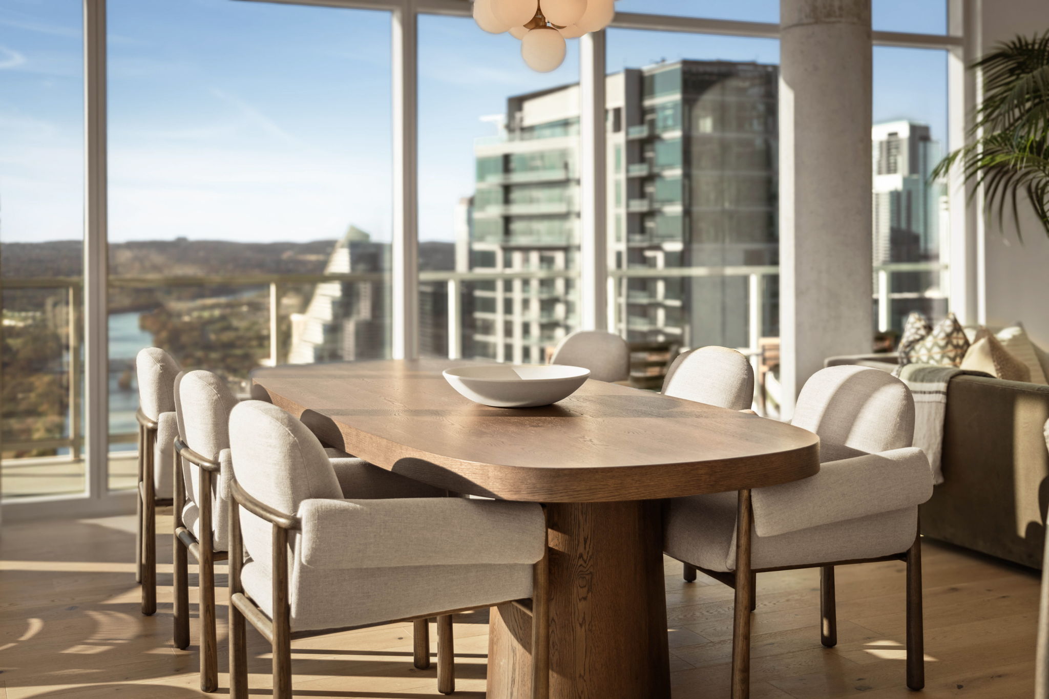 A modern dining area in the Paseo Penthouse features a large wooden table surrounded by eight upholstered chairs. Floor-to-ceiling windows reveal a cityscape and river view, allowing natural light to fill the room.