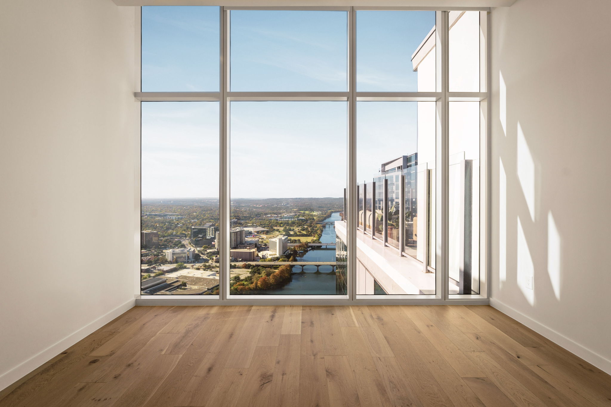 Empty room with light wood floor, white walls, and large floor-to-ceiling windows offering a stunning paseo penthouse cityscape view with a river, bridge, and buildings under a clear blue sky.